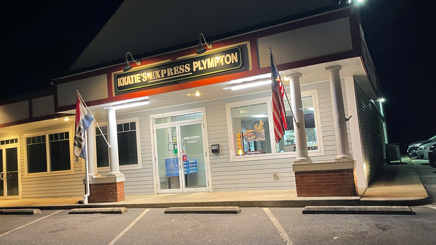 Exterior of a KKatie's Express Plympton, at night. Features American flags and white exterior with brick accents.