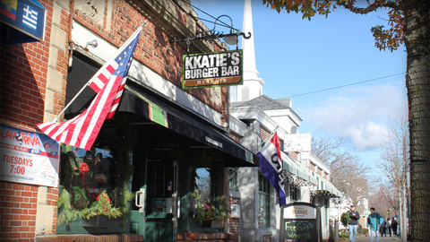 KKatie's Burger Bar with American flag. Brick building, black awning, sunny day, sidewalk with people.