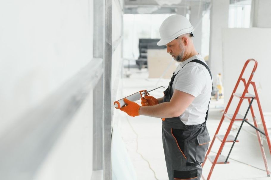 Construction worker using a power tool on a white wall in a bright hallway, with a ladder nearby