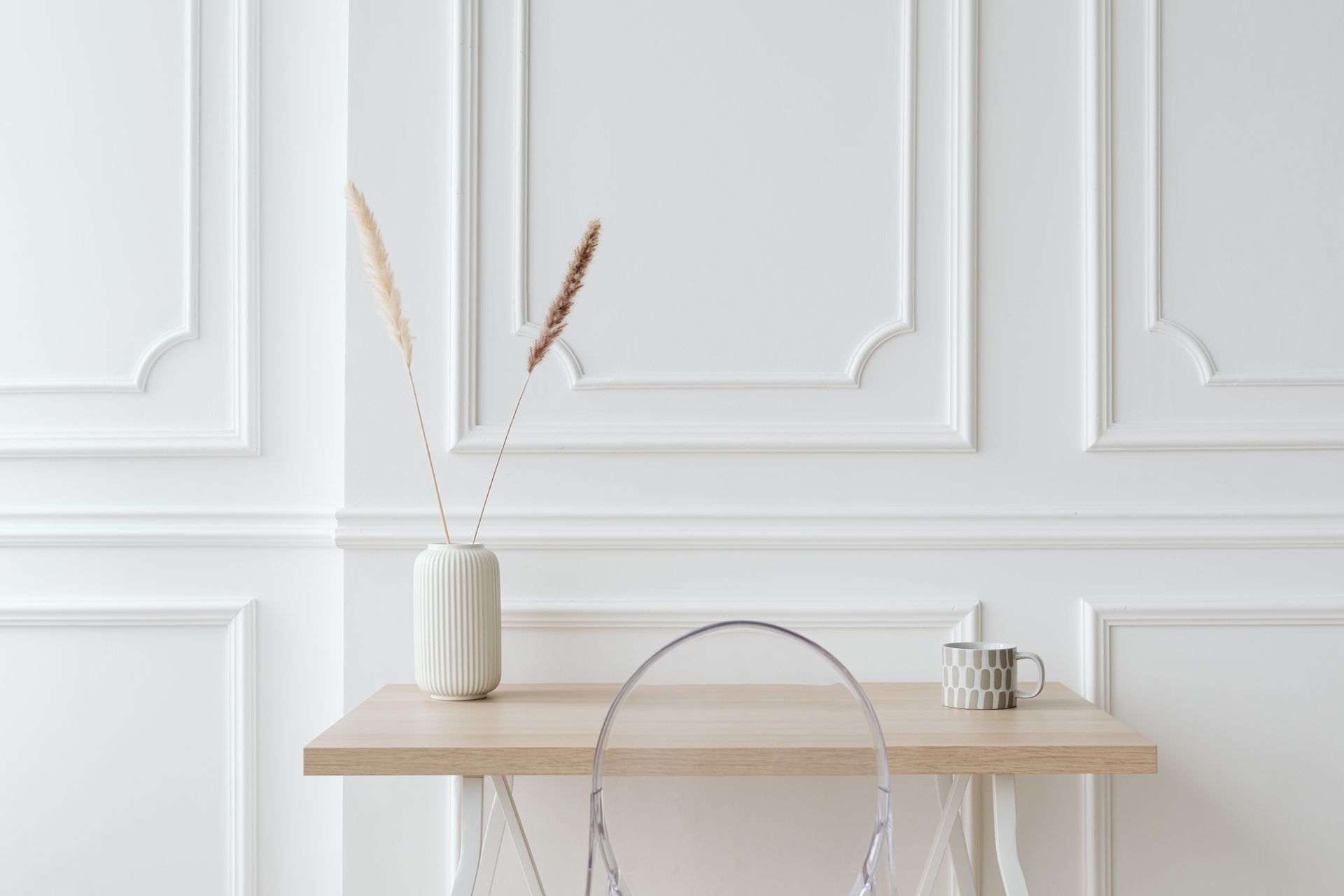 Minimalist white wall with a light wood table, vase with dried stems, and a clear chair.