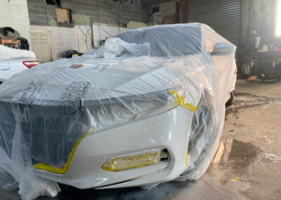 White car covered in plastic sheeting and tape, likely in a garage, ready for painting or repair.