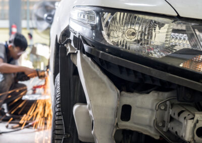 Car being repaired in auto shop, sparks flying, front bumper detached.