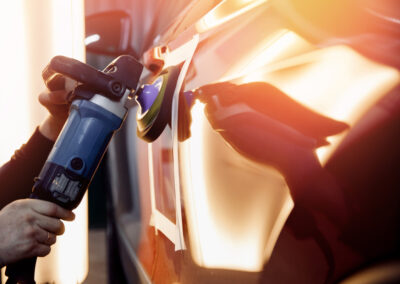 Person using a power buffer to polish a car's surface, likely for detailing or paint correction, with bright sunlight.