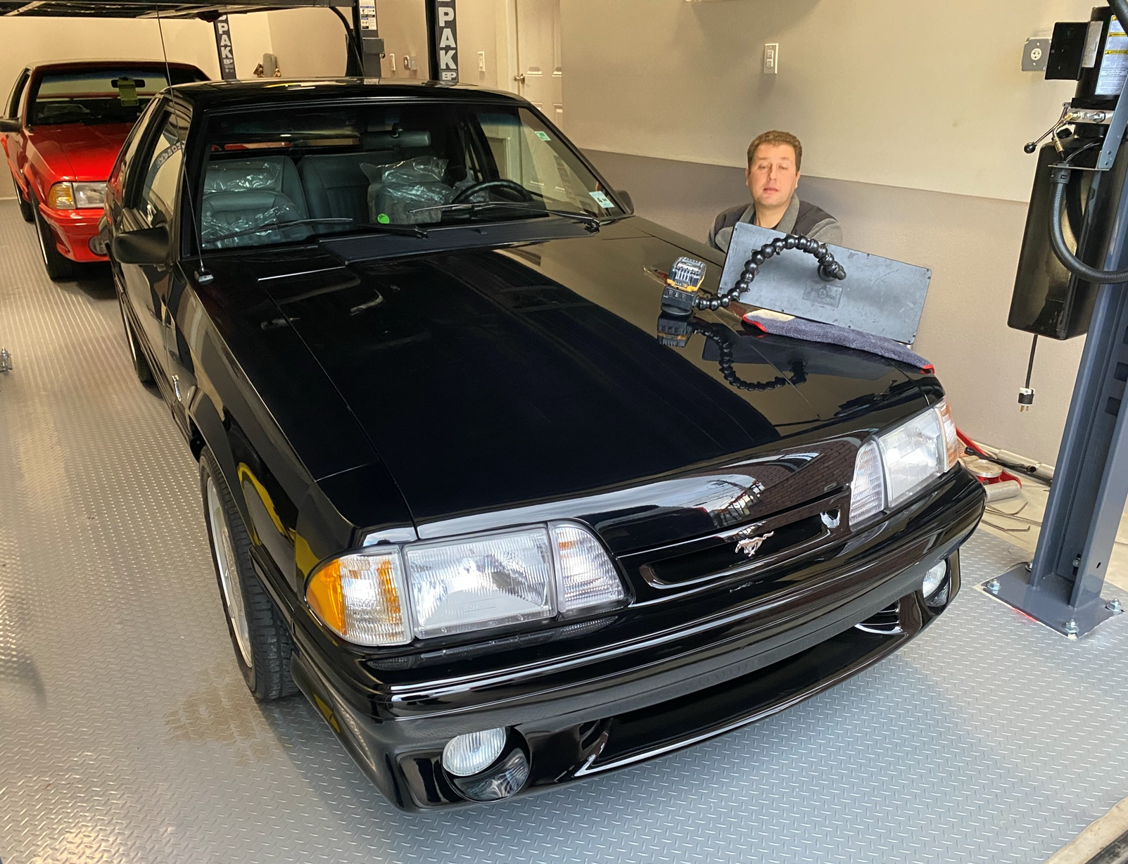Black Ford Mustang in a garage; man stands near the hood, smiling. Red Mustang visible behind it.