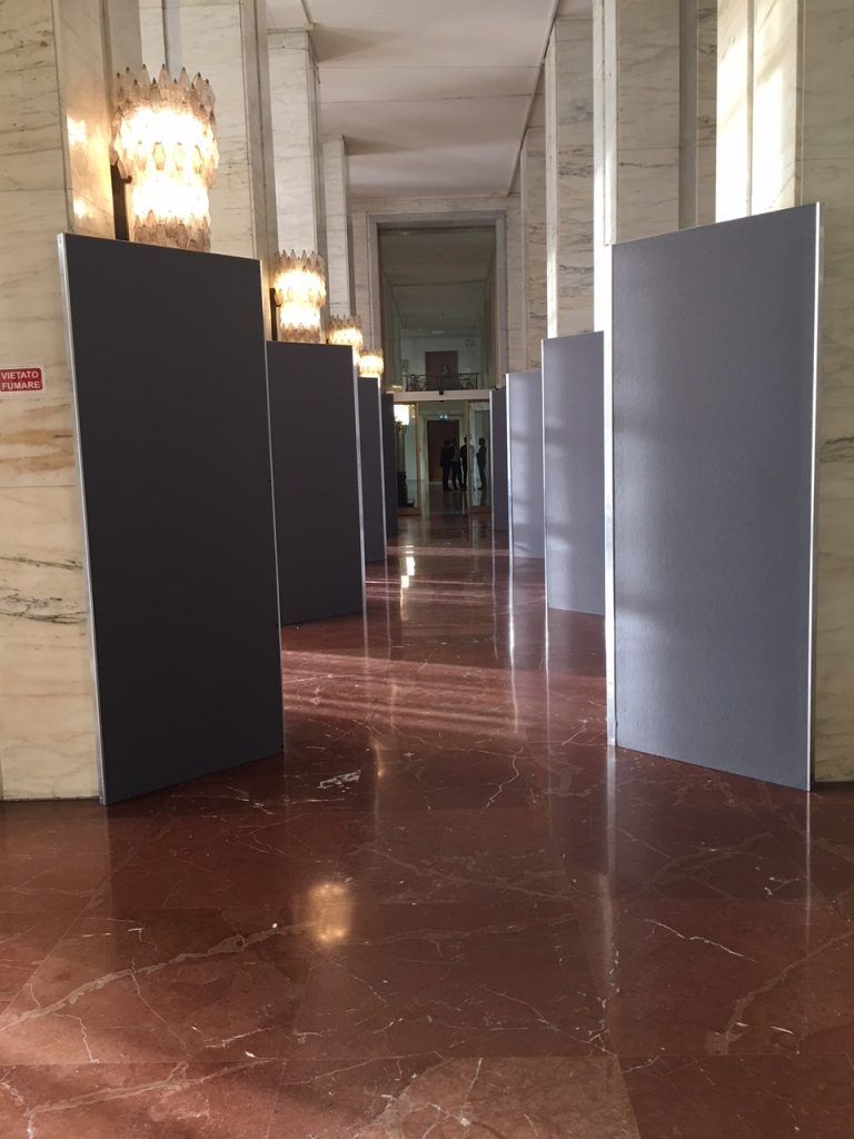 Lungo corridoio della galleria con pavimento in marmo e alti pannelli espositivi neri e grigi.