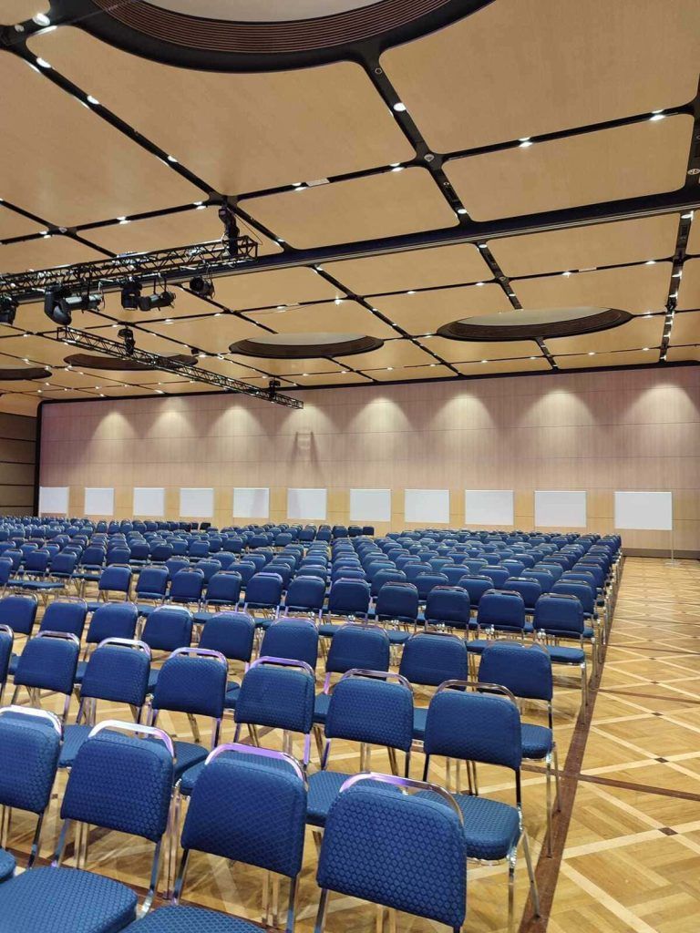 Sala conferenze vuota con file di sedie blu e soffitto rivestito in legno.