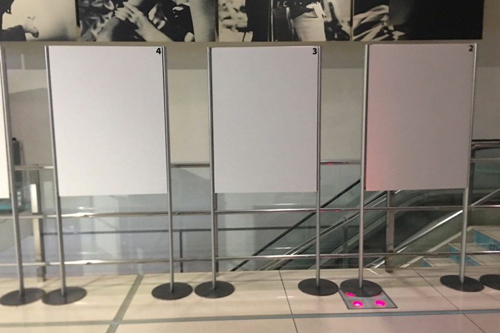 In una stanza simile a una galleria, tre pannelli espositivi vuoti su dei supporti, con foto in bianco e nero appese sopra.