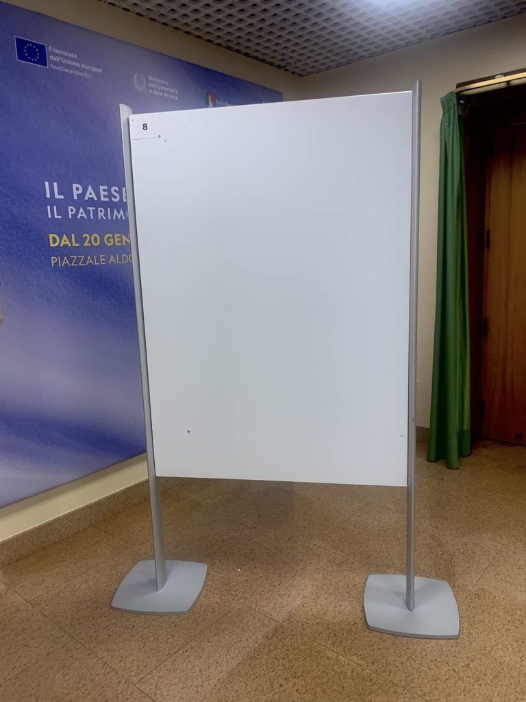 Pannello espositivo bianco autoportante su gambe metalliche in un corridoio con parete blu e tenda verde.