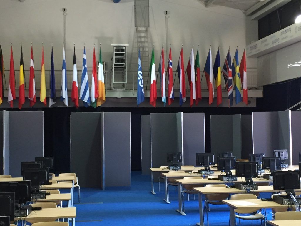 Sala conferenze vuota con file di tavoli e monitor sotto una fila di bandiere colorate.