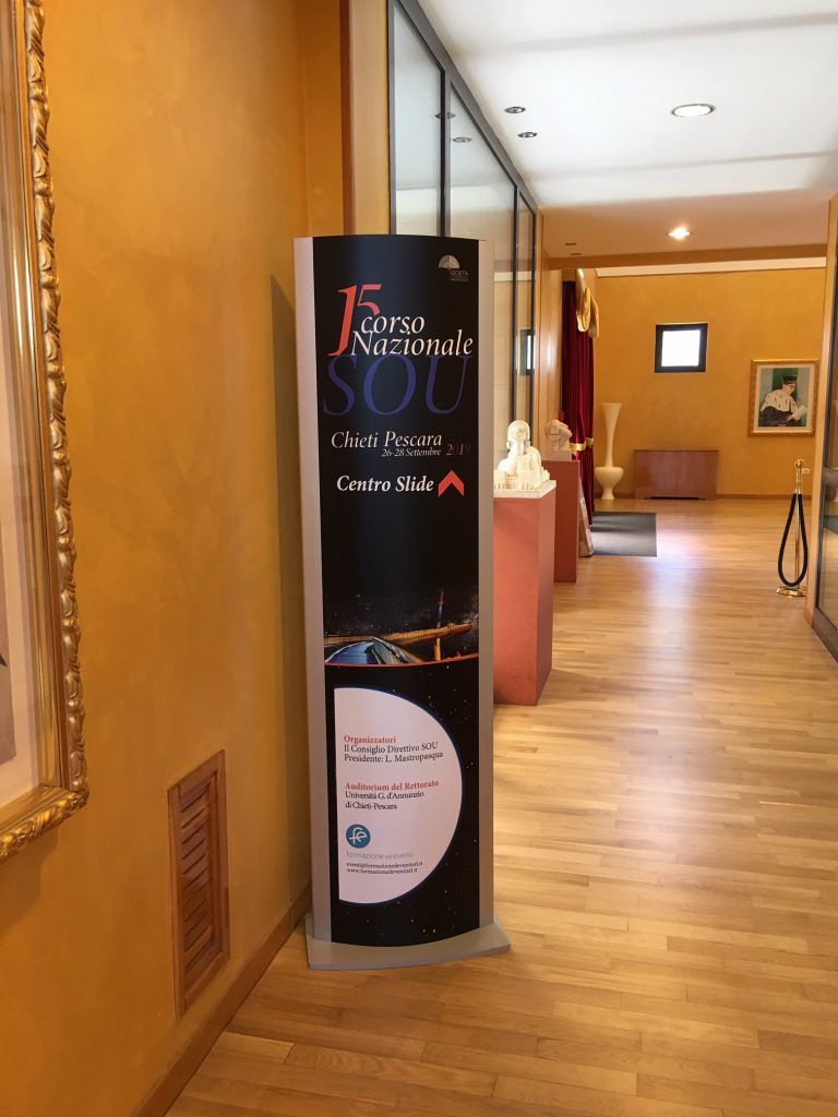 Atrio con un'alta insegna nera per eventi accanto al banco della reception, in un corridoio con pavimento in legno.