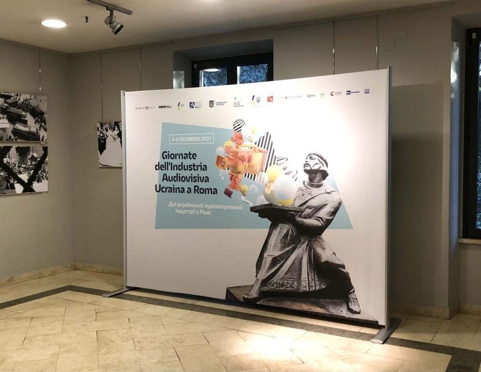 Esposizione nella hall con un grande poster bianco, grafica colorata e una figura in bianco e nero che tiene in mano dei fiori e un bambino