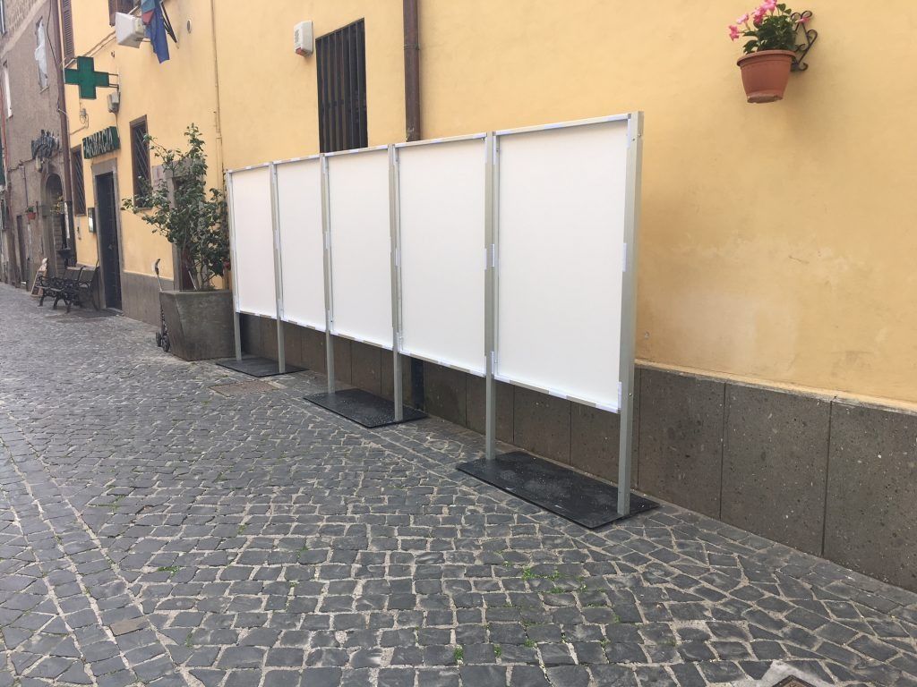 Delle pareti divisorie bianche per esterni fiancheggiano una stretta strada acciottolata accanto a un edificio giallo con vasi di fiori.