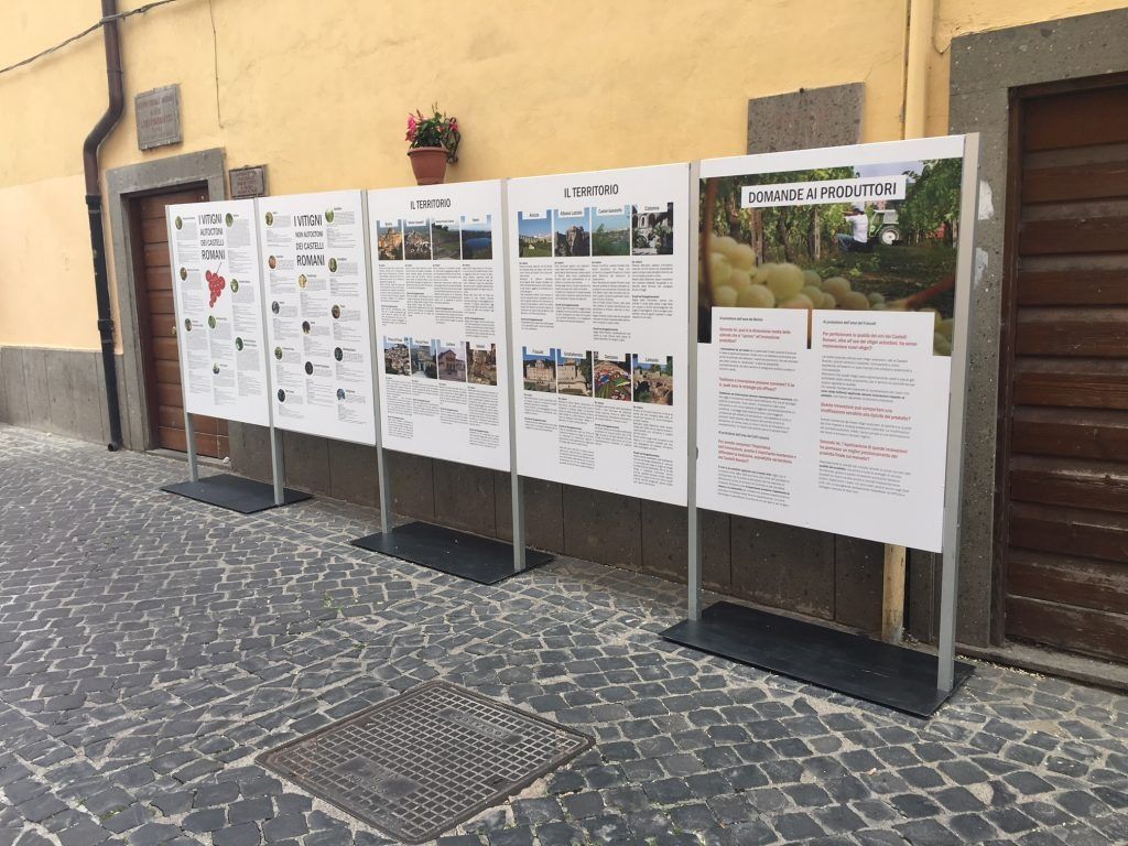 Pannelli espositivi esterni con poster su un muro giallo in una strada acciottolata