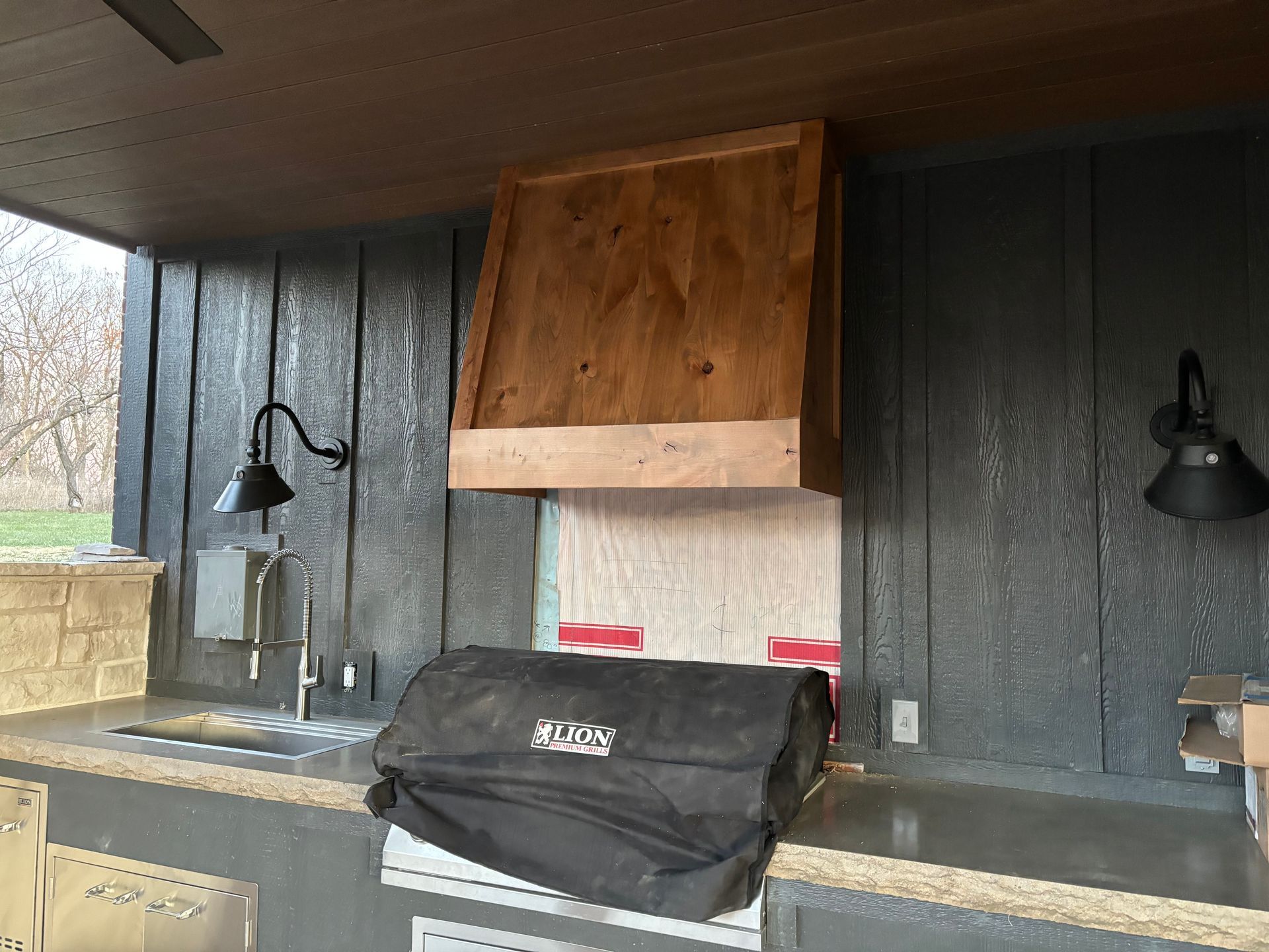 Outdoor kitchen with wooden hood over a grill, gray countertop, black wall sconces.
