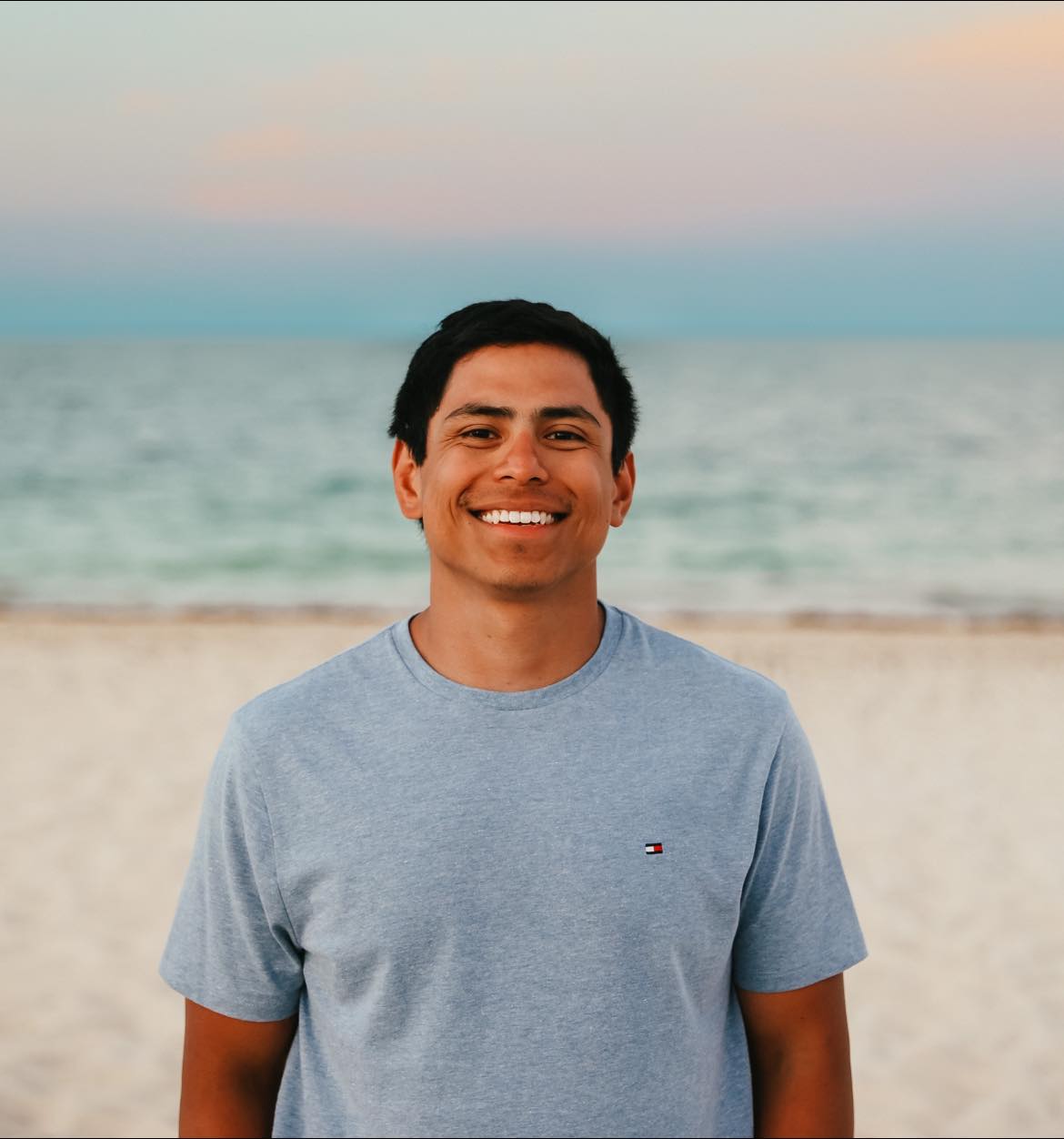 Man smiling on a beach, wearing a gray t-shirt. Ocean and sunset in background.