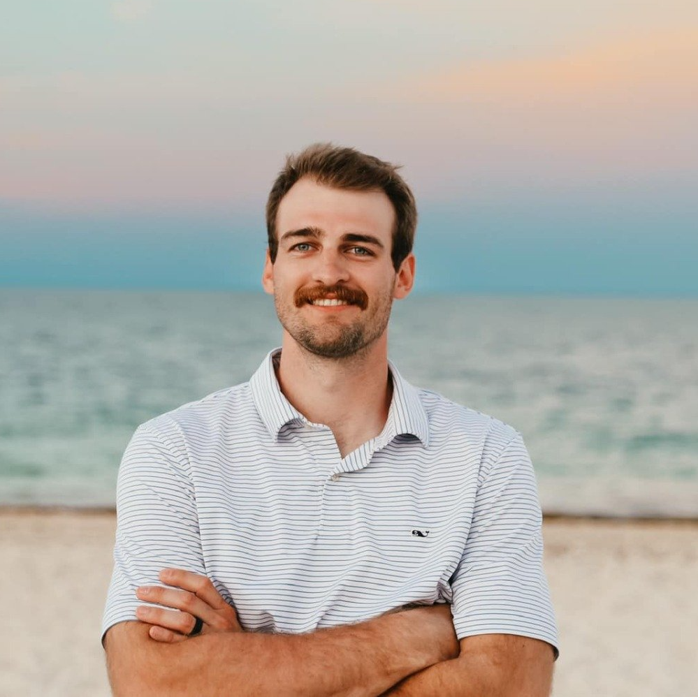Man with a mustache, arms crossed, smiles at the camera on a beach with the ocean and a colorful sky in the background.