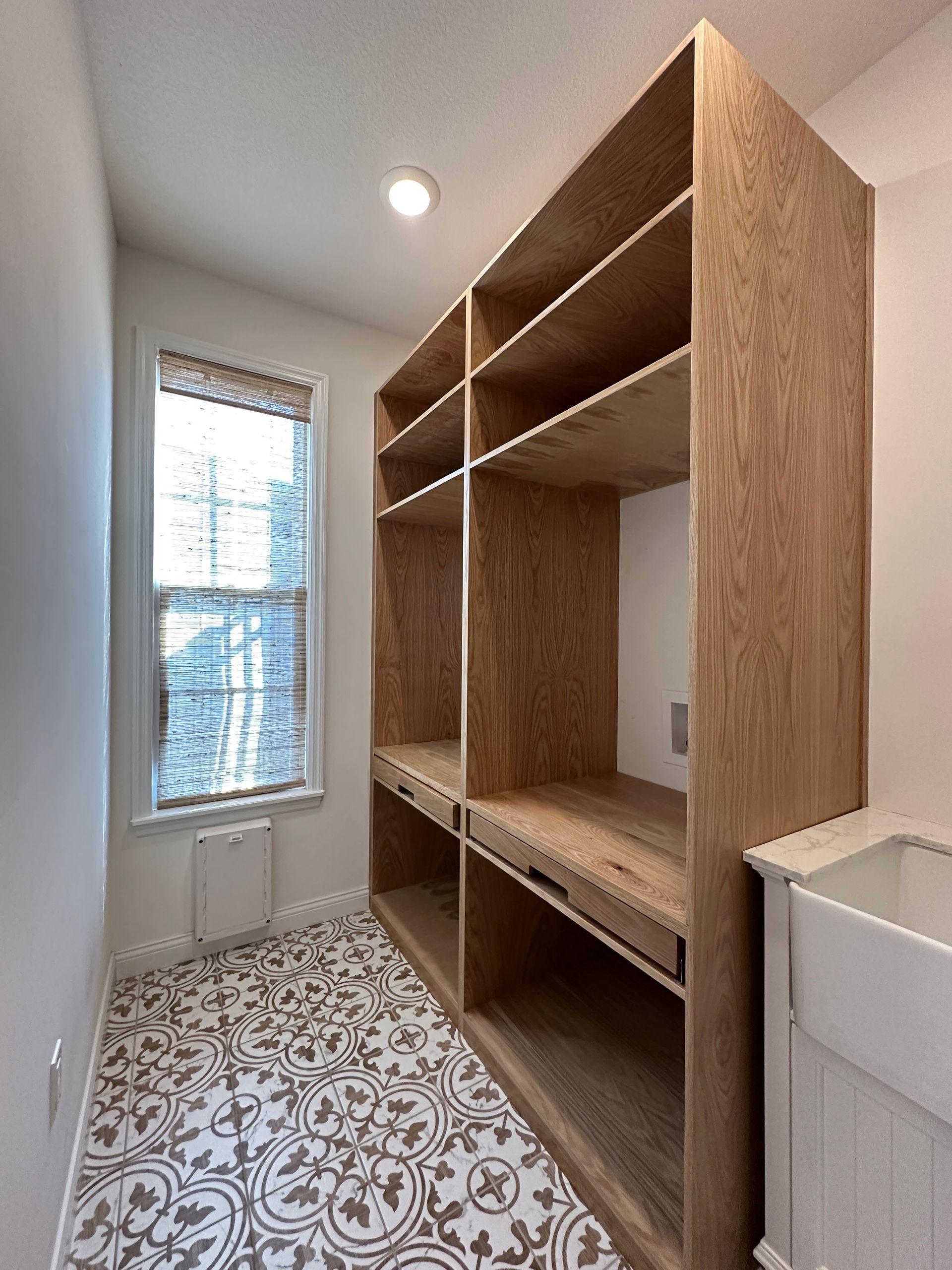 Laundry room with built-in wooden shelving unit, white sink, patterned floor, and window with shade.