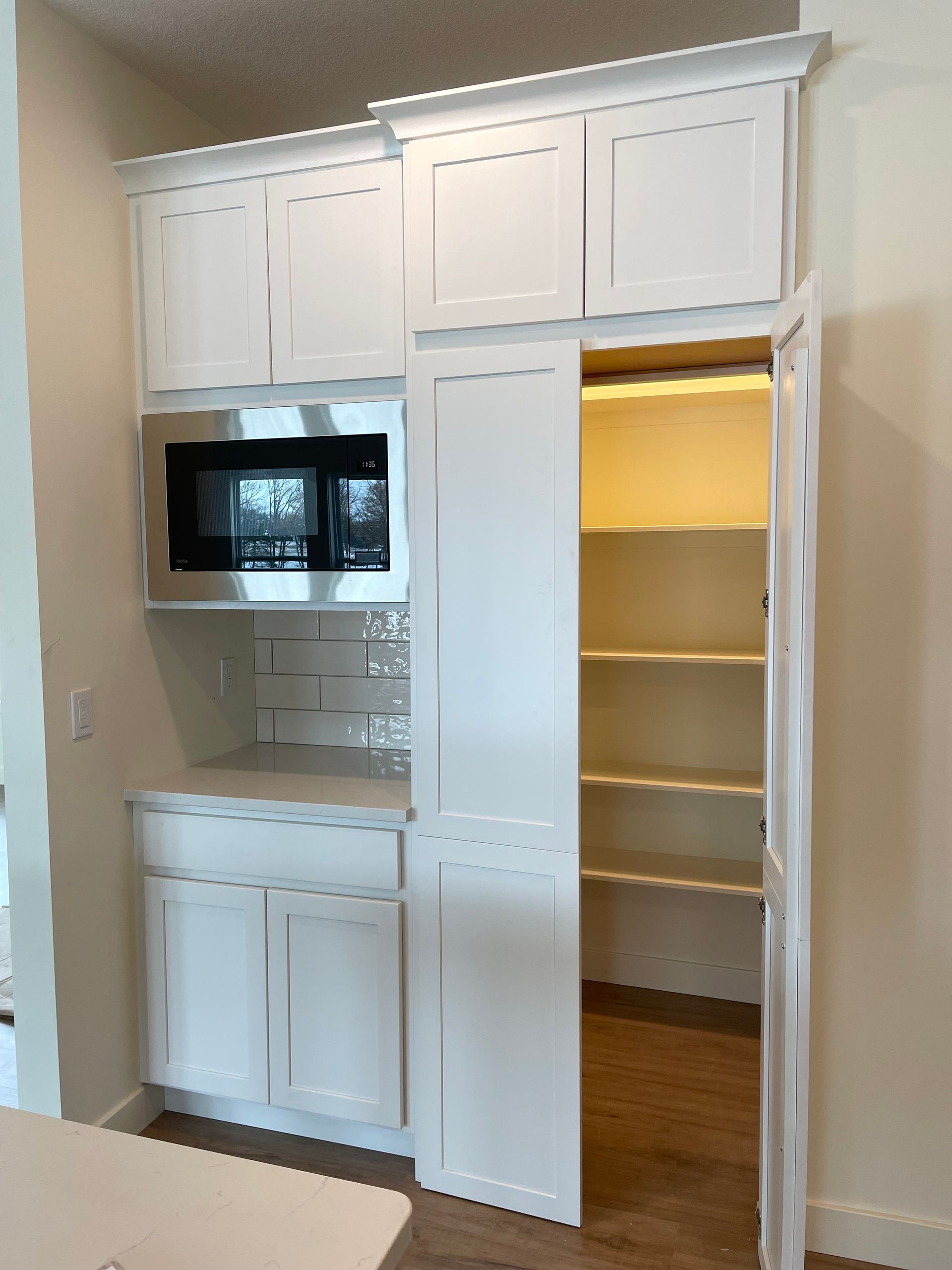 White built-in kitchen cabinets with microwave and pantry. Pantry door is open, shelves visible, on wood floor.