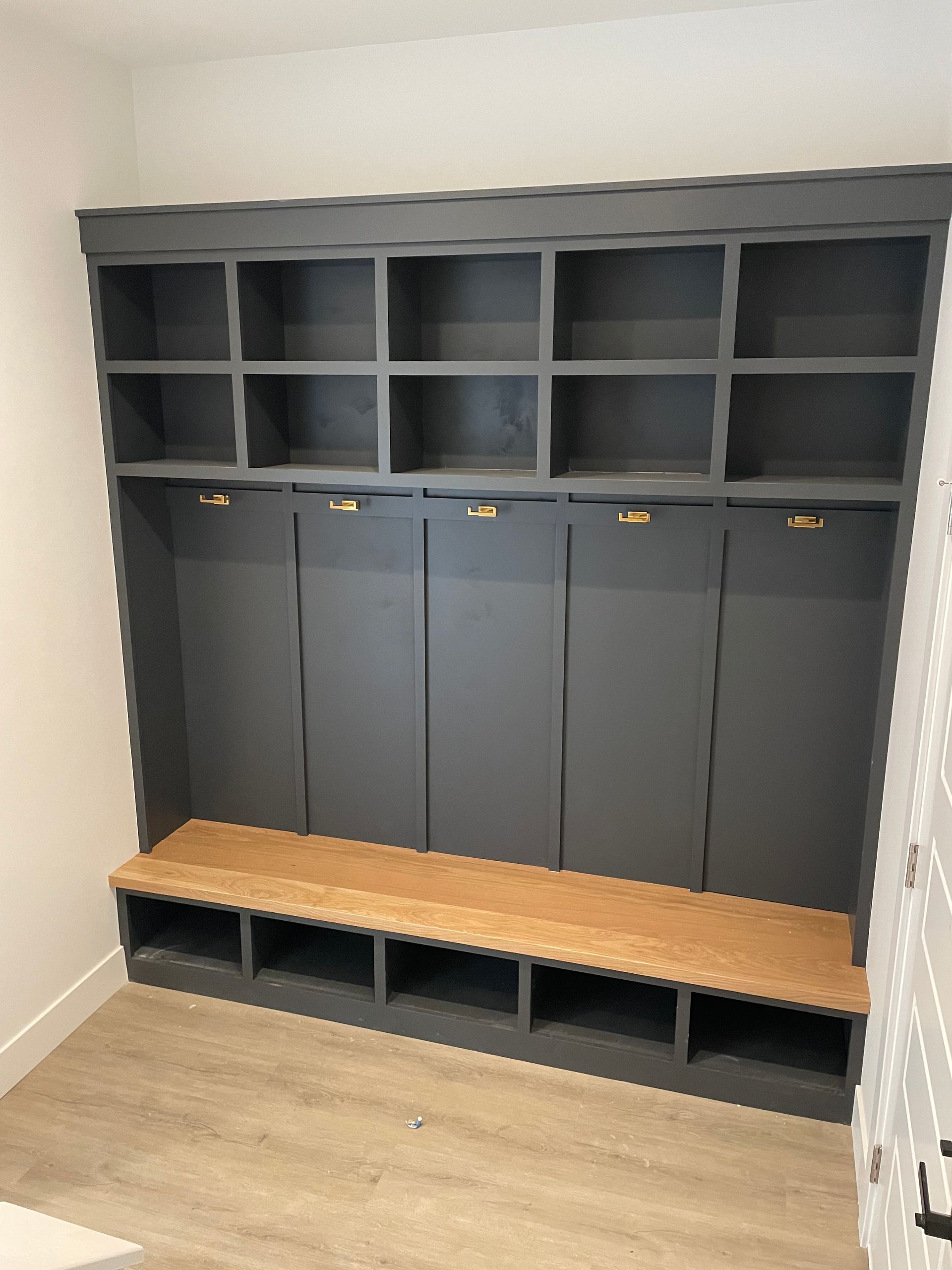 Gray built-in mudroom storage unit with cubbies, hooks, and bench, on light wood-look floor.