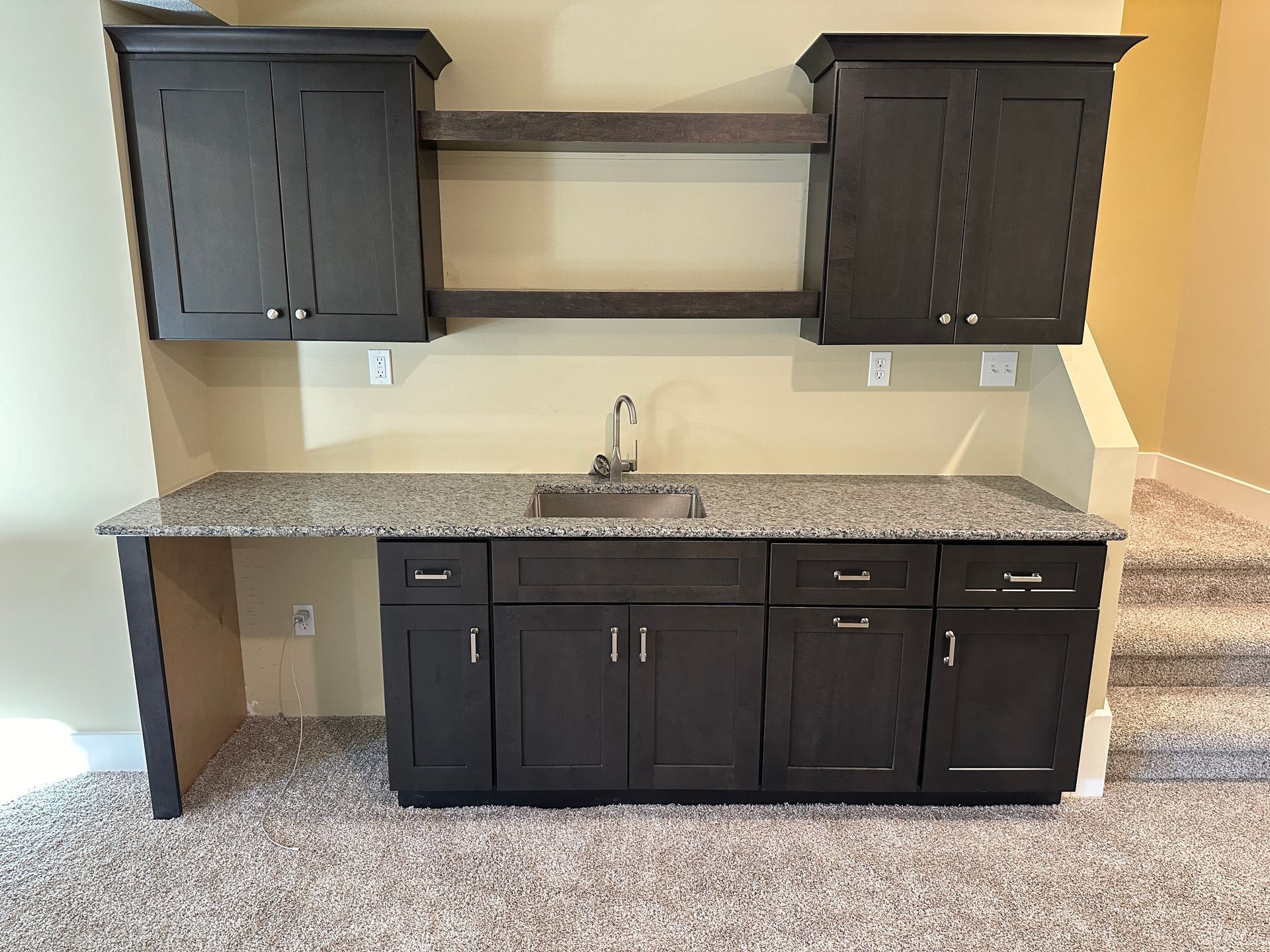 Dark cabinets and granite countertop bar area with sink, shelving, and stair access.