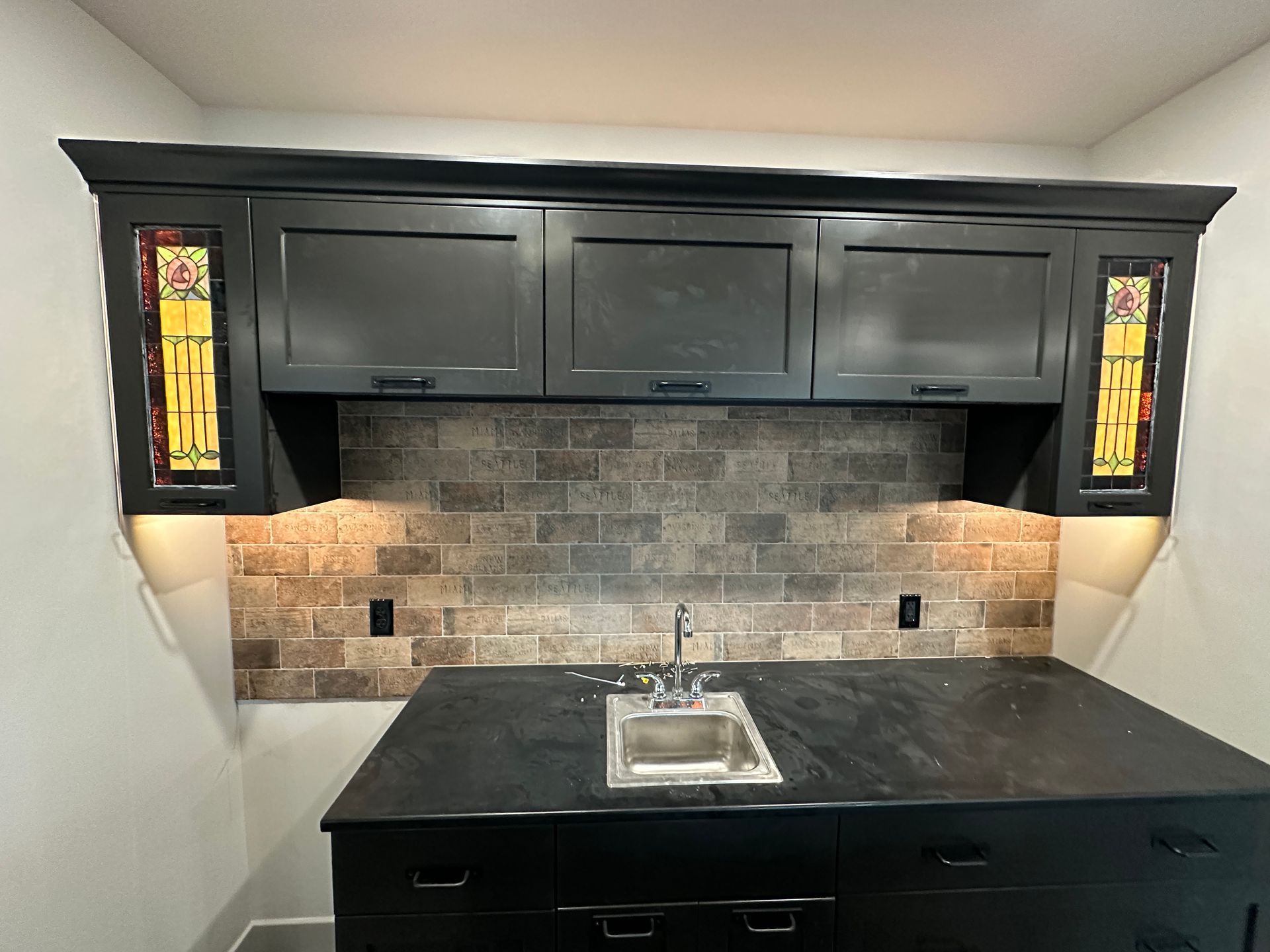 Black cabinets with stained glass accents above a dark countertop island with a sink, against a brick backsplash.