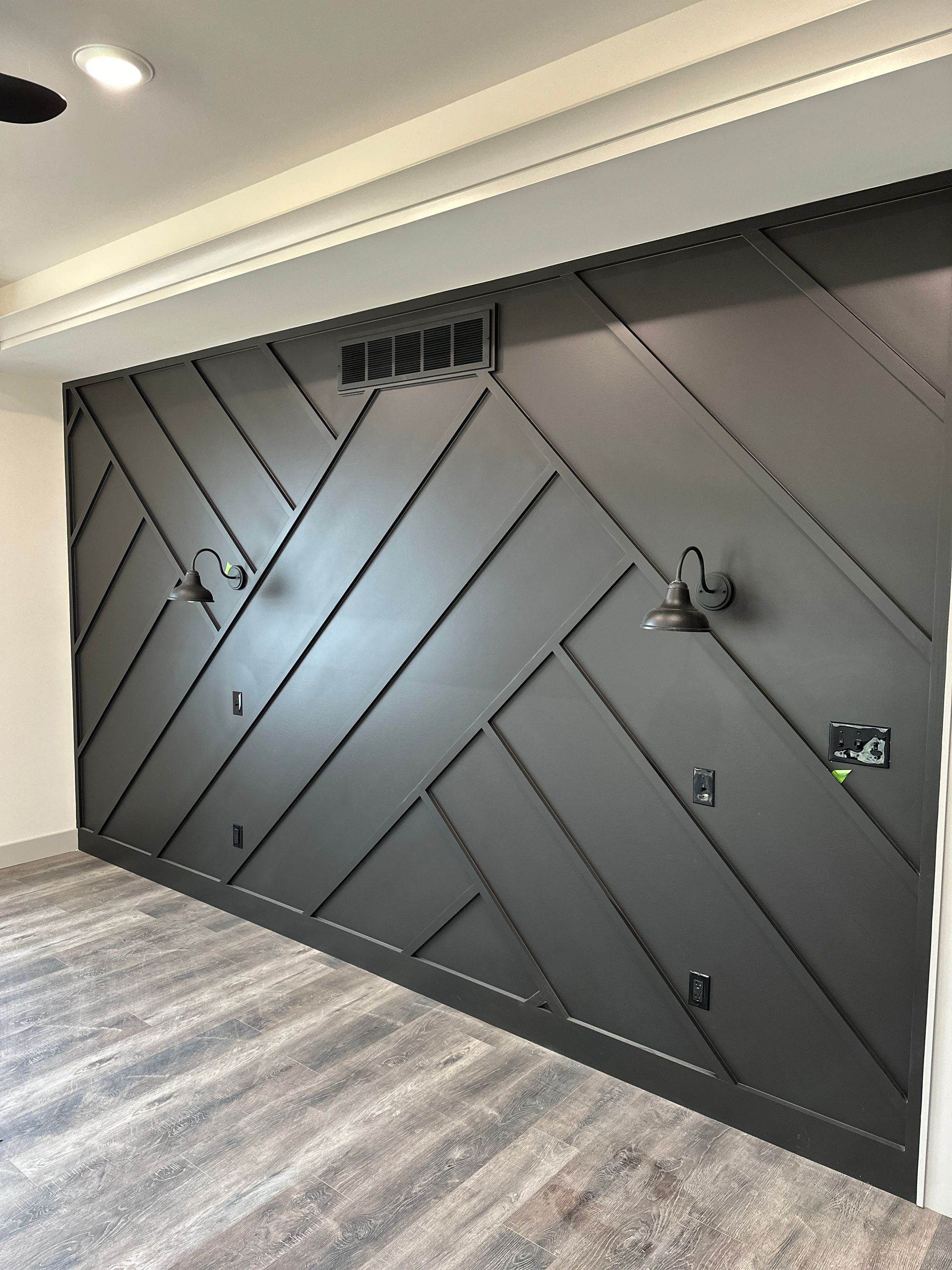Dark gray accent wall with herringbone pattern, sconces, and air vent; installed above wood-look flooring.