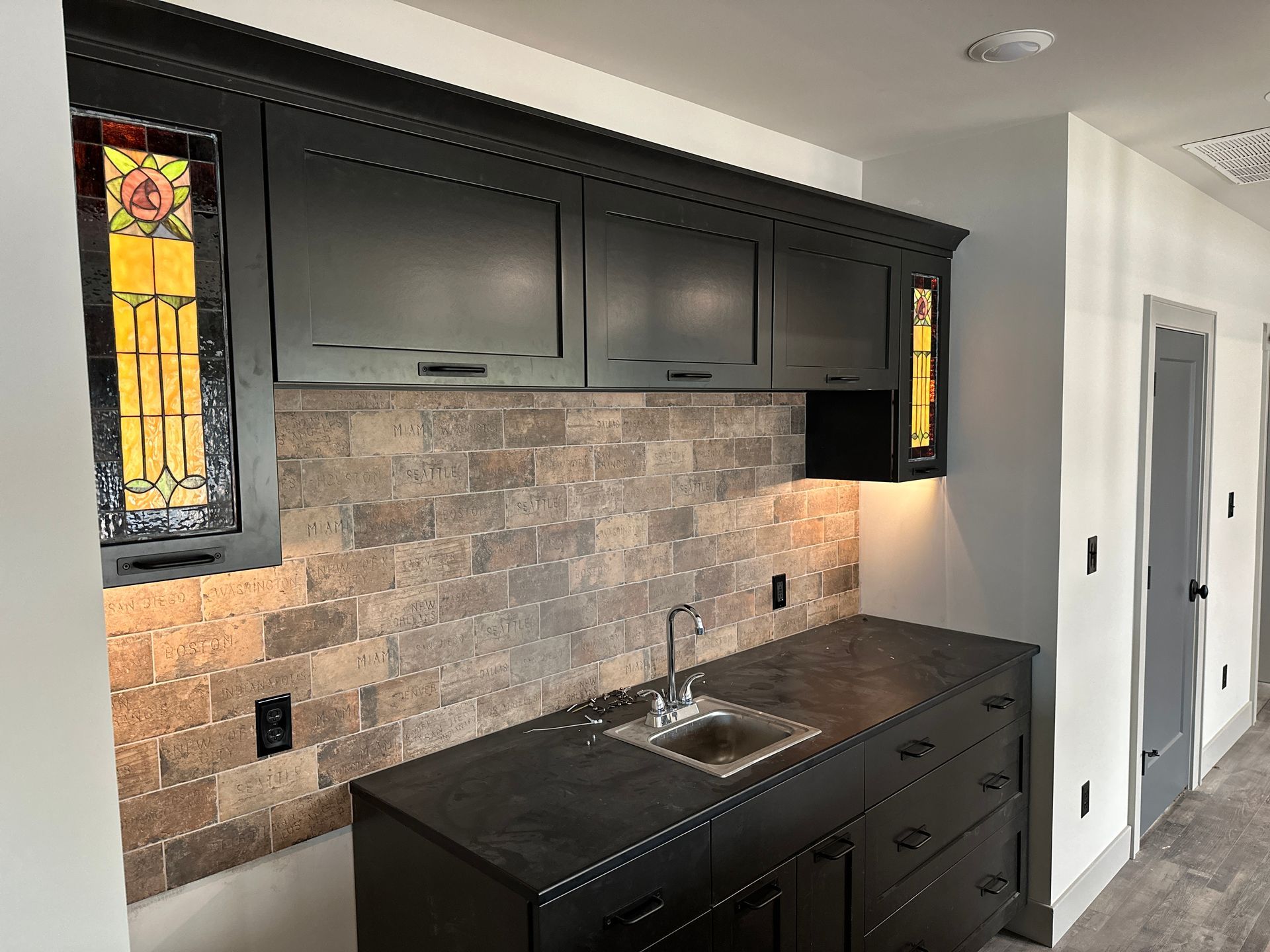 Black cabinetry with a stone backsplash and a sink. Stained glass window.
