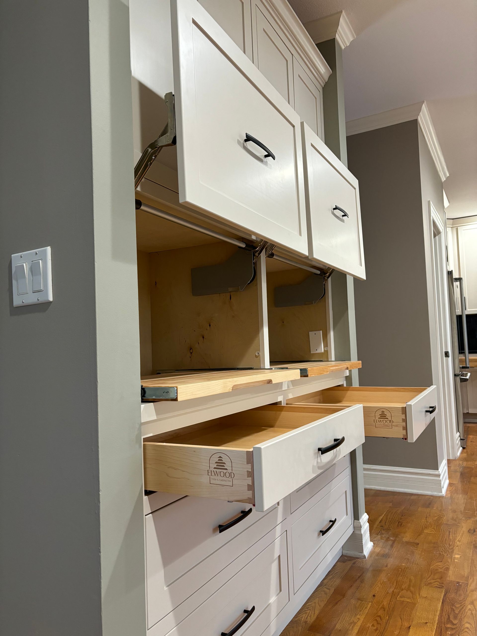 White kitchen cabinets, some open, revealing shelves and drawers. Gray wall and wood floor.