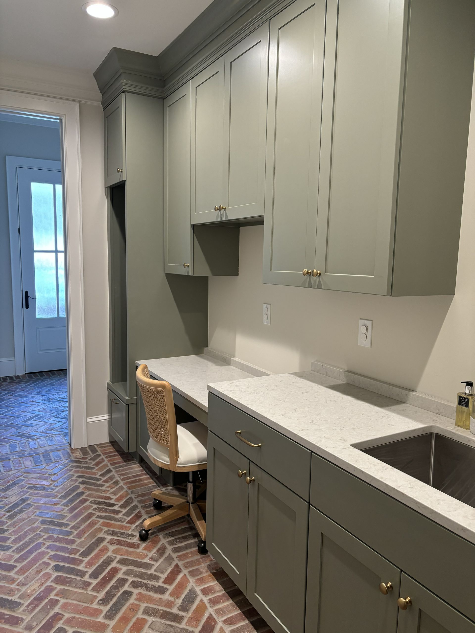 Green cabinetry and white countertop workspace in a room with a brick floor.