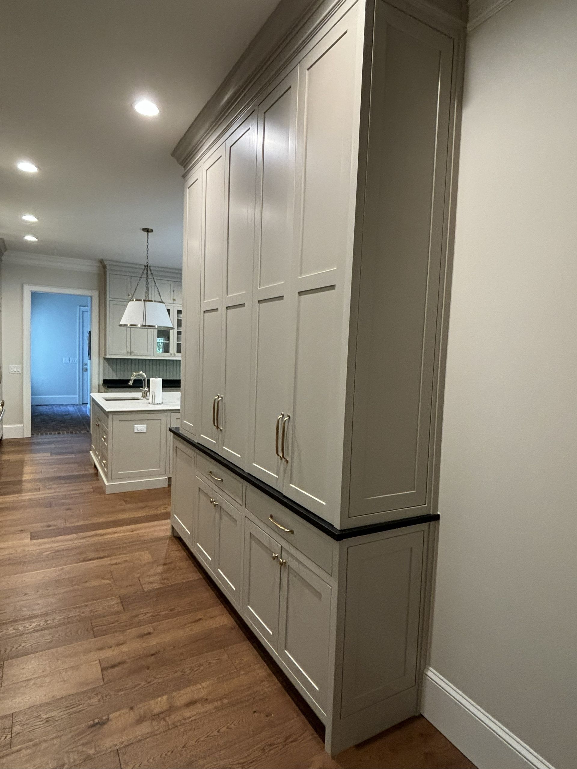 Tall, light gray kitchen cabinets against a beige wall, next to wood flooring. A kitchen is in the background.