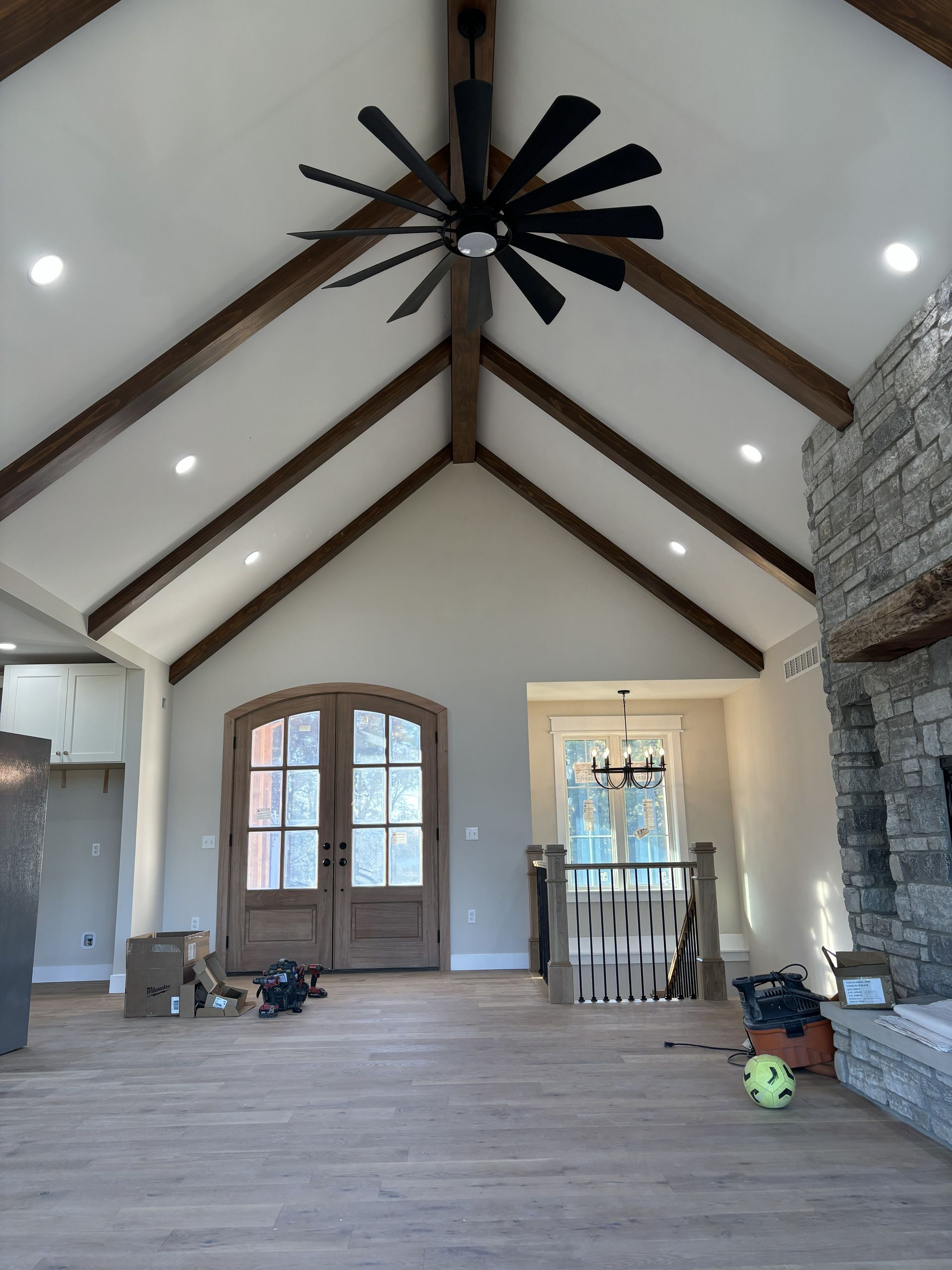 Spacious room with vaulted ceiling, wood beams, and large fan. Wood flooring, double doors, stone fireplace.