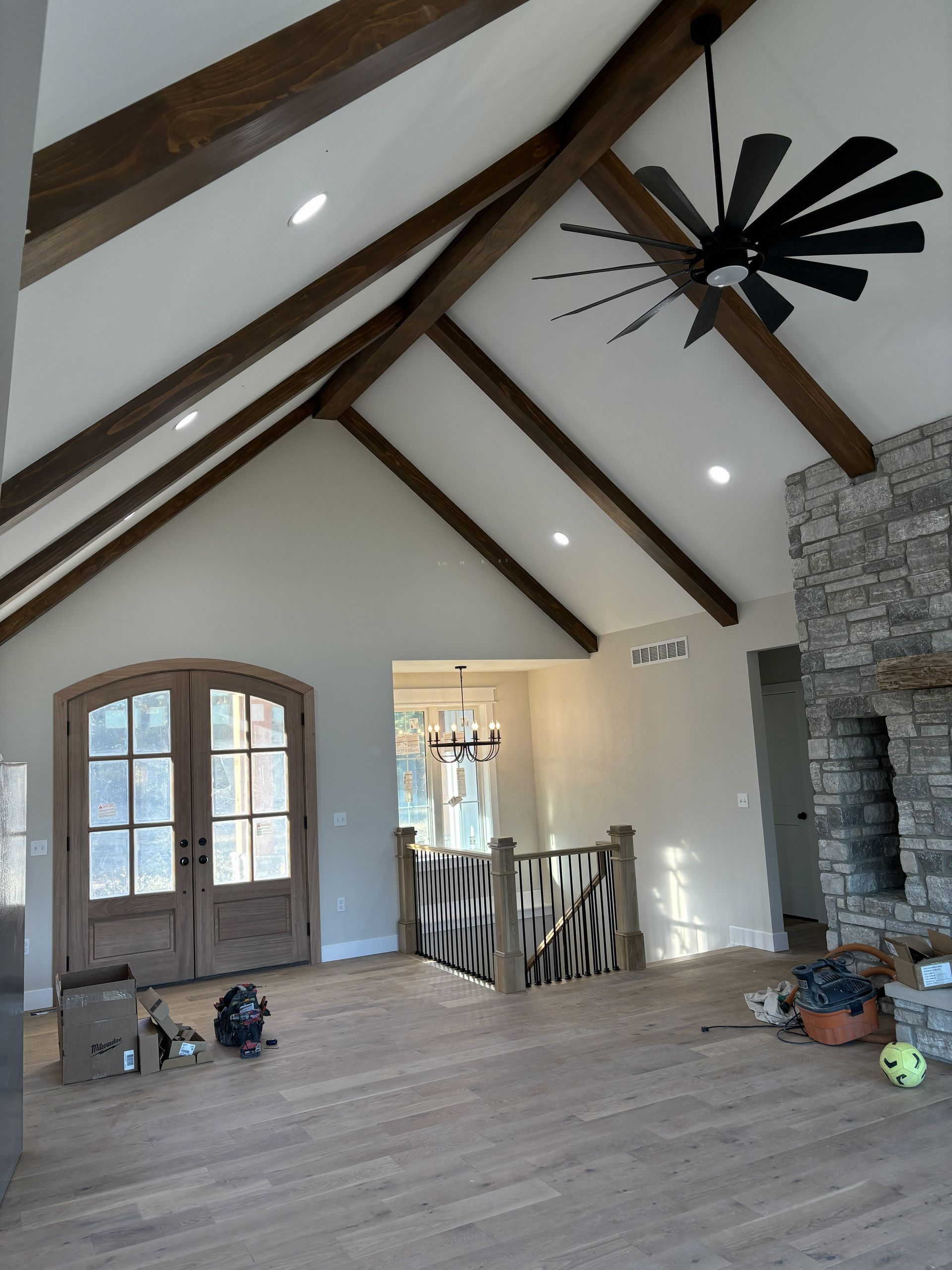 Vaulted ceiling living room with wooden beams and a stone fireplace. Double doors, hardwood floors, and a black ceiling fan.