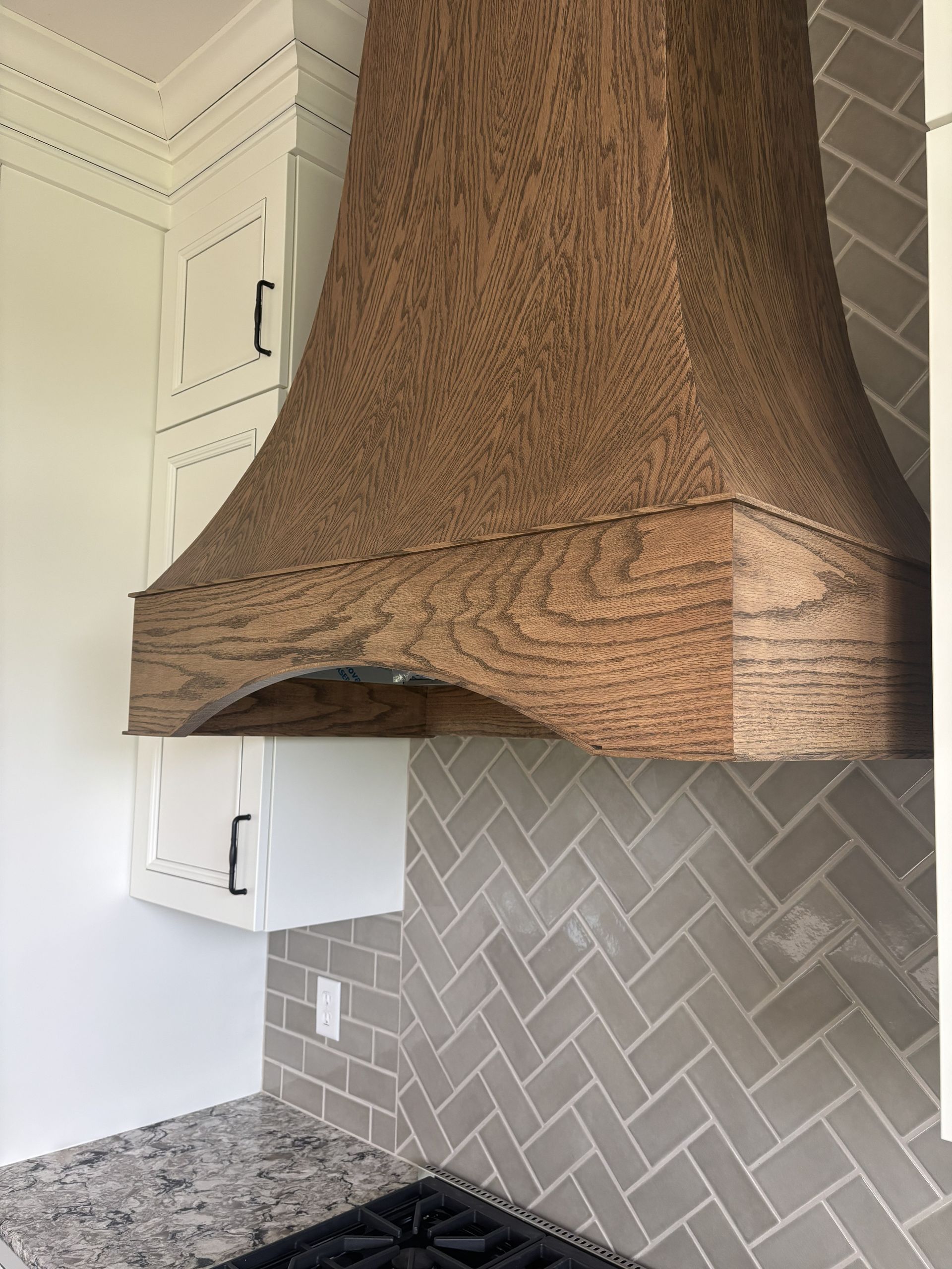 Wood range hood above gray herringbone tile backsplash and gas range.