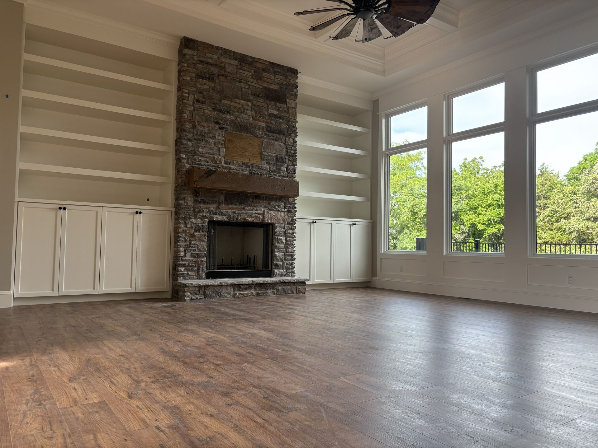 Empty living room with fireplace, built-in shelves, large windows, and dark wood floors.