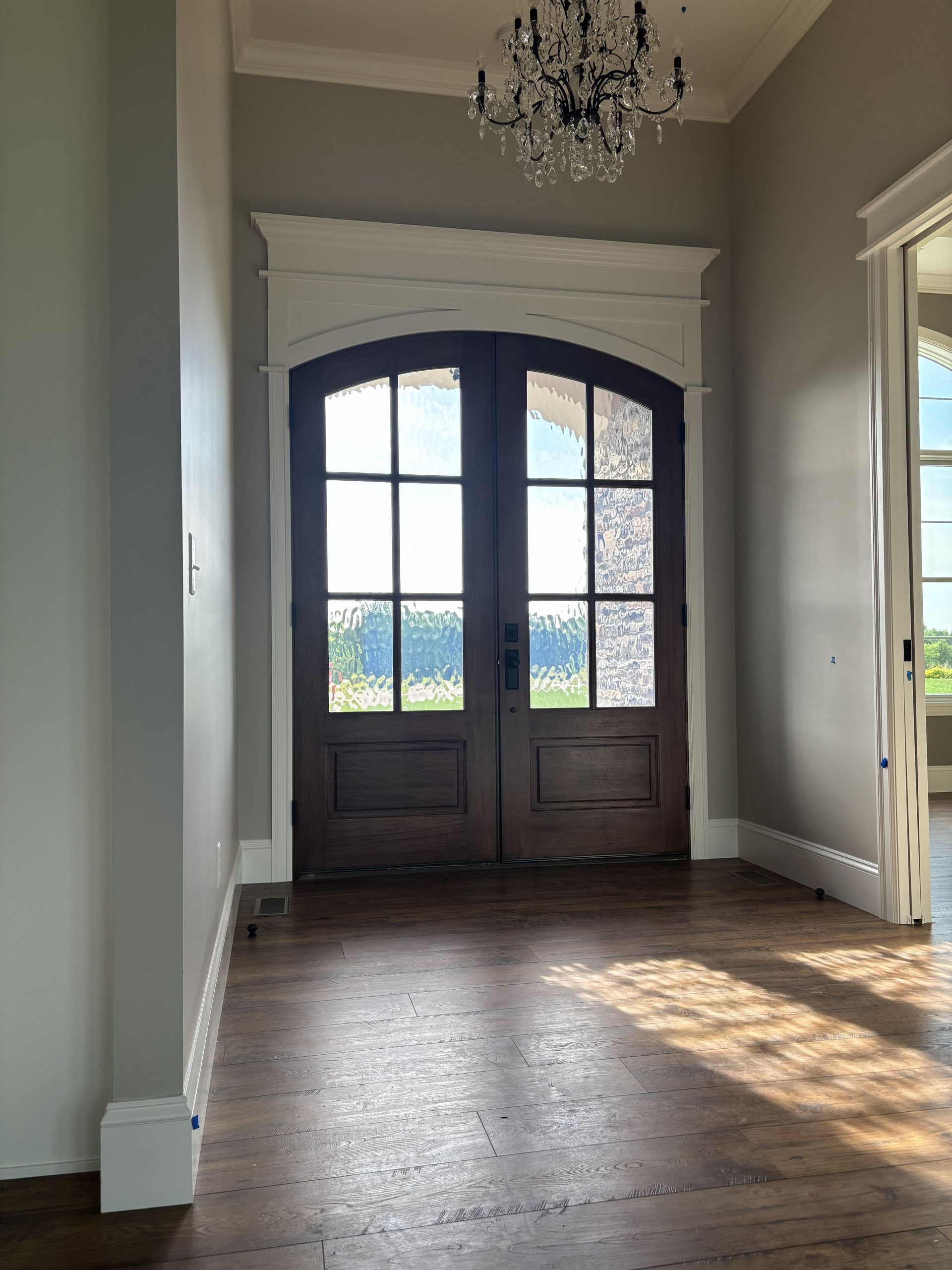 Dark wooden double doors with arched tops, viewed from inside. White trim, chandelier, and a hallway with wood floor.