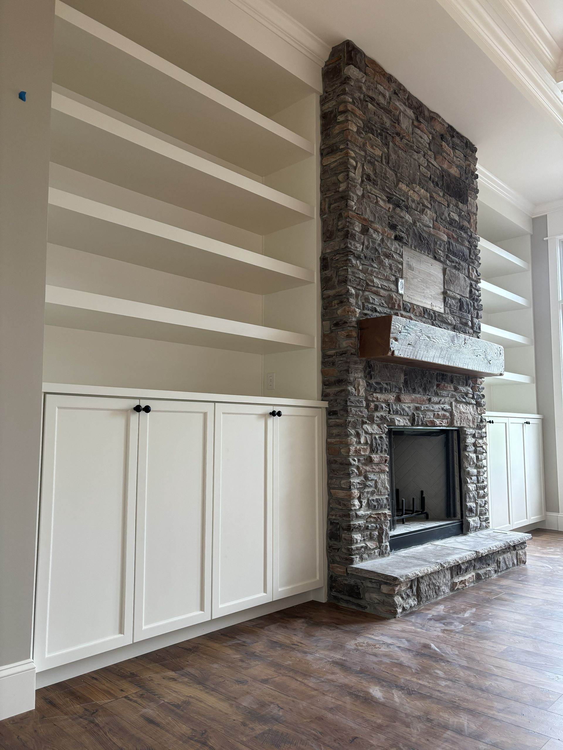 Built-in white bookshelves flank a stone fireplace with a wooden mantel in a room with hardwood floors.