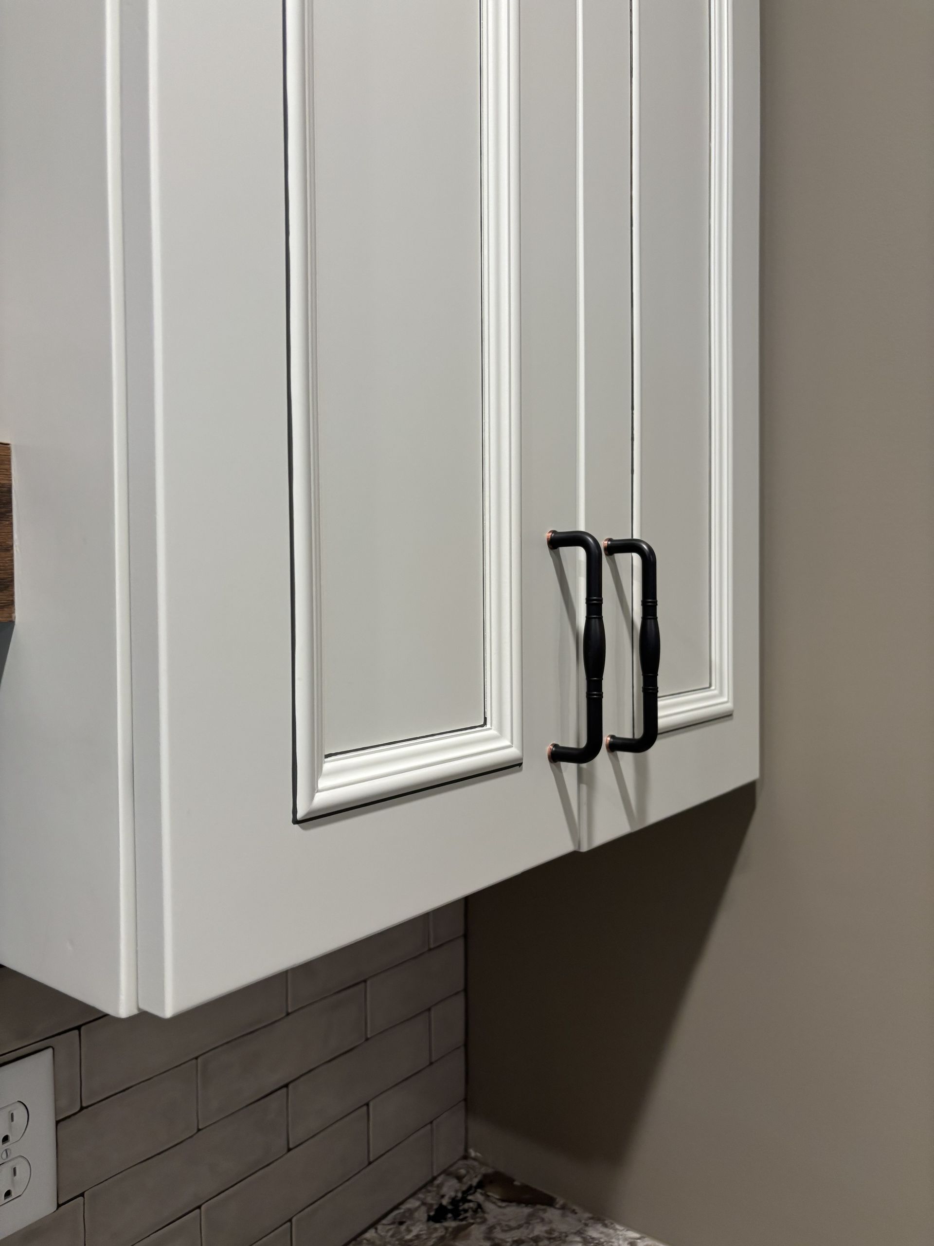 Gray kitchen cabinet with black handles, set against a neutral wall and brick-style backsplash.