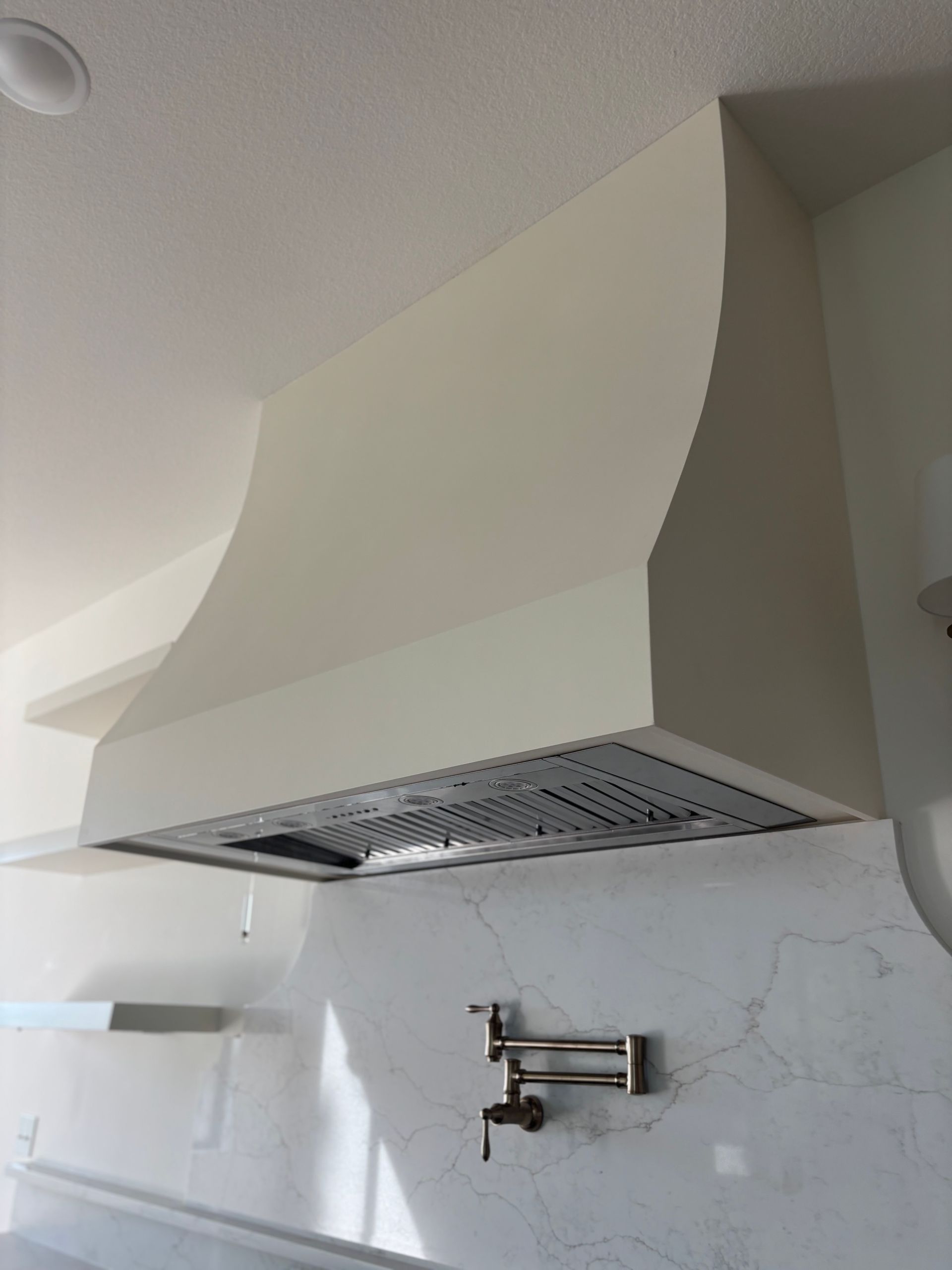 Cream-colored kitchen range hood with curved sides over a stainless steel range and white marble backsplash.
