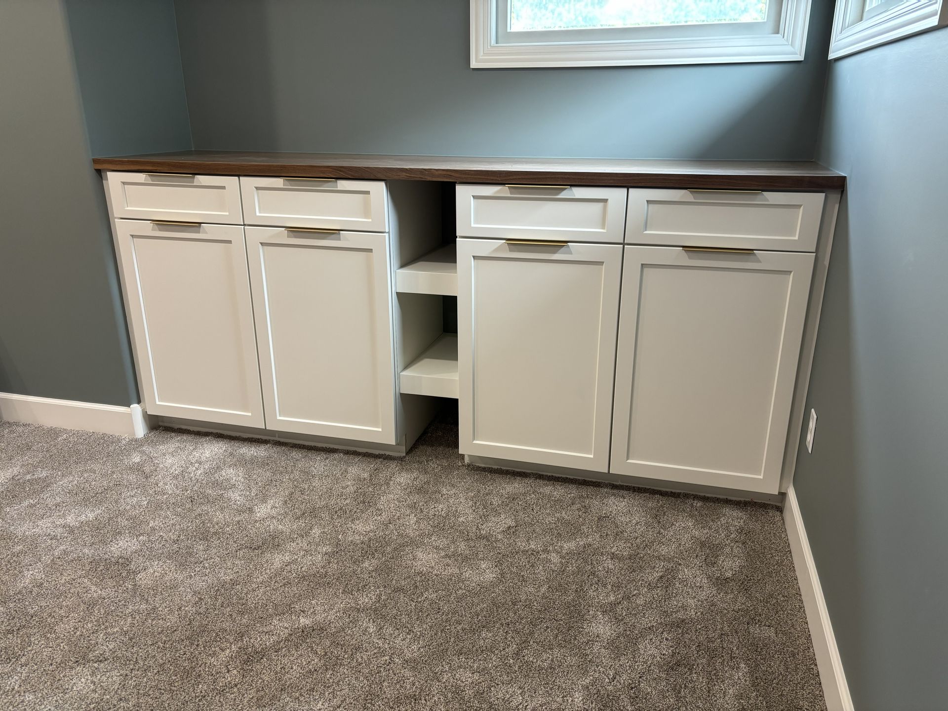 Built-in white cabinets with a brown countertop in a corner of a room with carpet and a window.