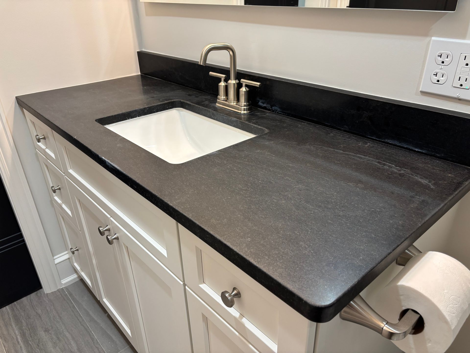 Bathroom vanity with dark countertop, white cabinet, and silver faucet. Toilet paper roll holder on side.