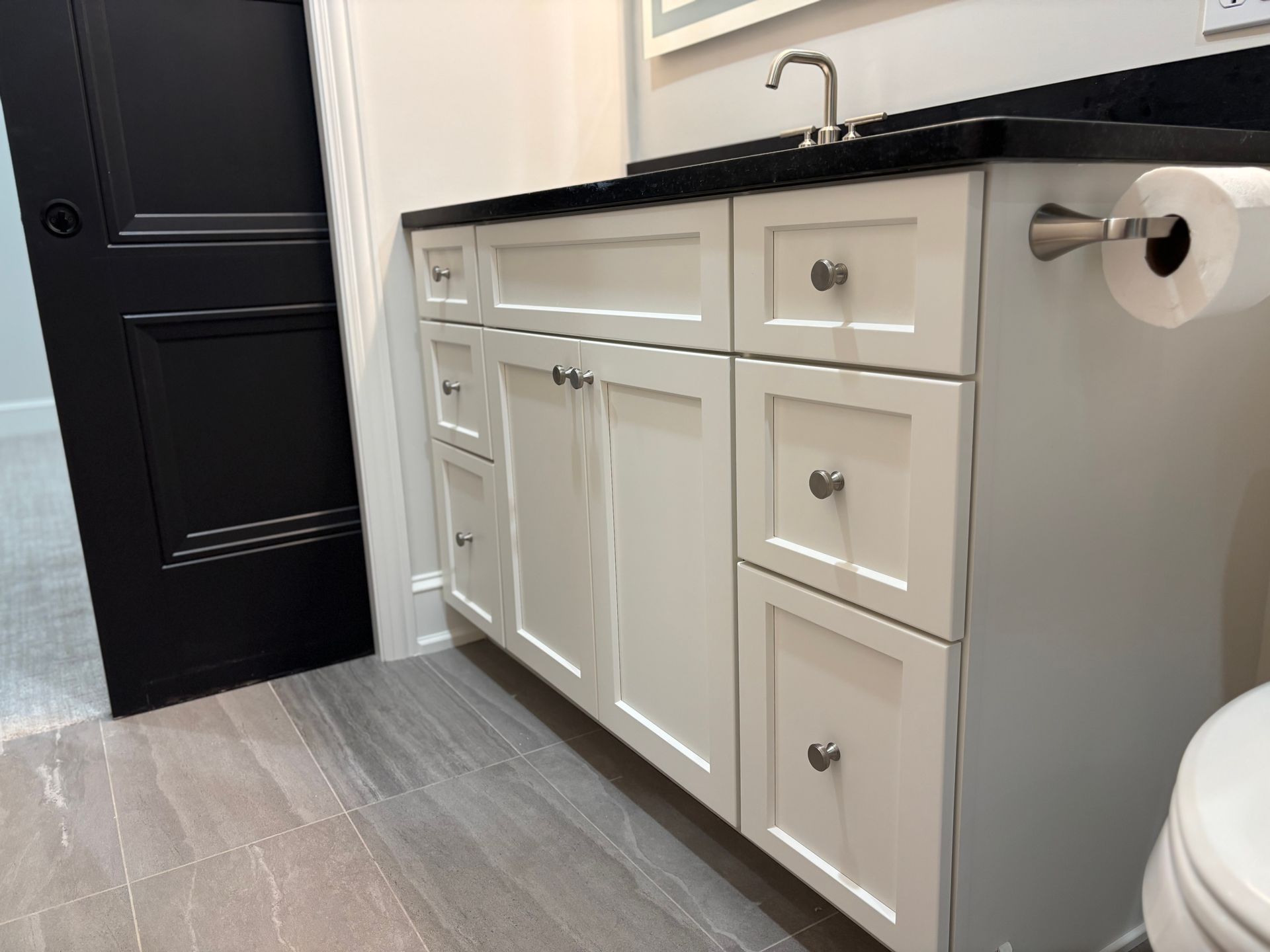 White bathroom vanity with black countertop, drawers, and black door, toilet paper roll.