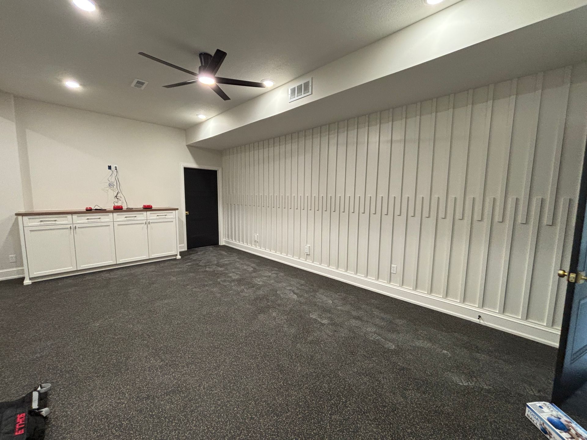 Empty room with white vertical paneling, dark ceiling fan, dark door, white cabinets, and dark speckled flooring.