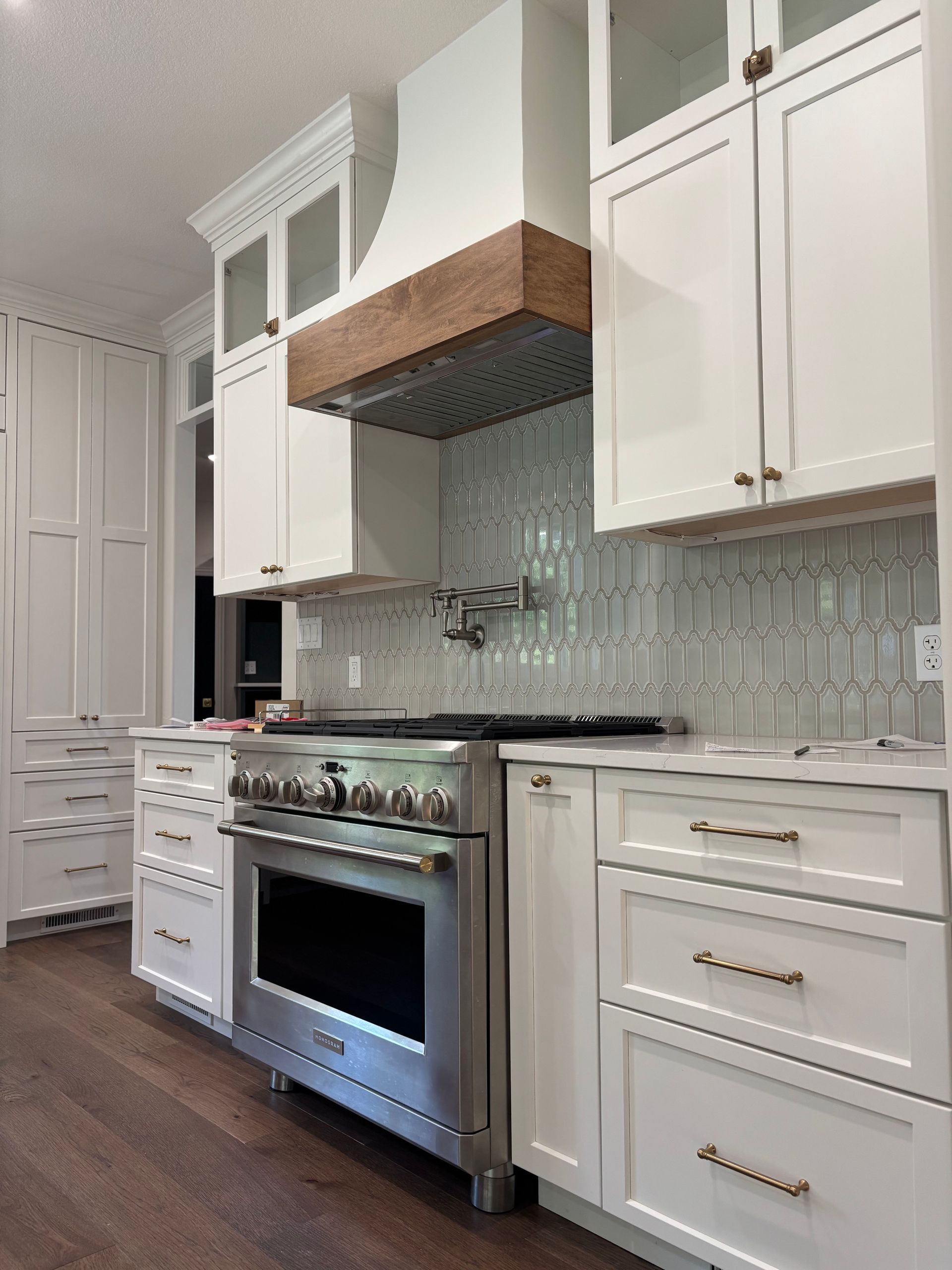 White kitchen with stainless steel oven, wood range hood, and light blue tile backsplash.