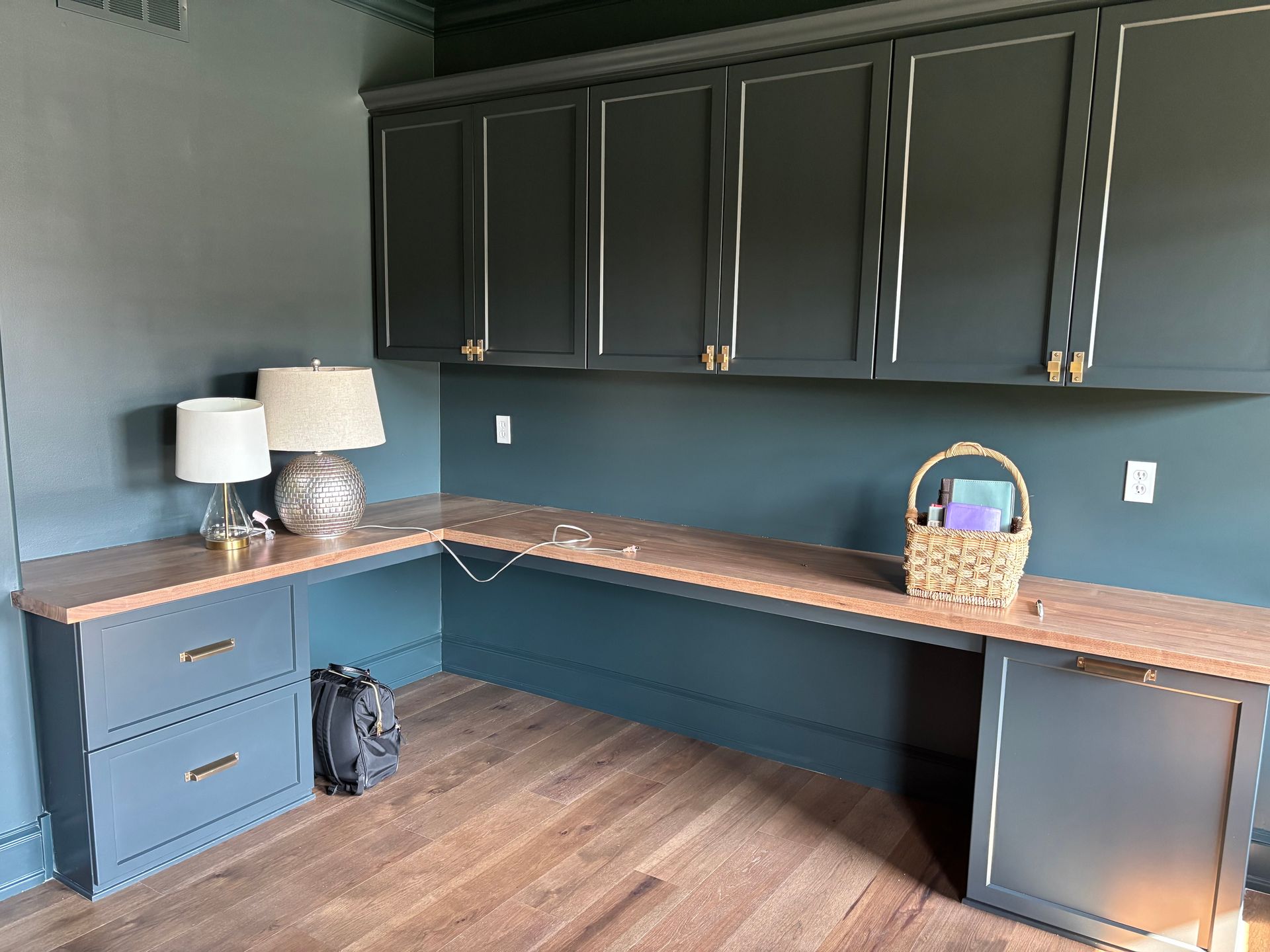 A teal home office with built-in desk and cabinets. Wooden desk, cabinets with gold hardware and lamps.