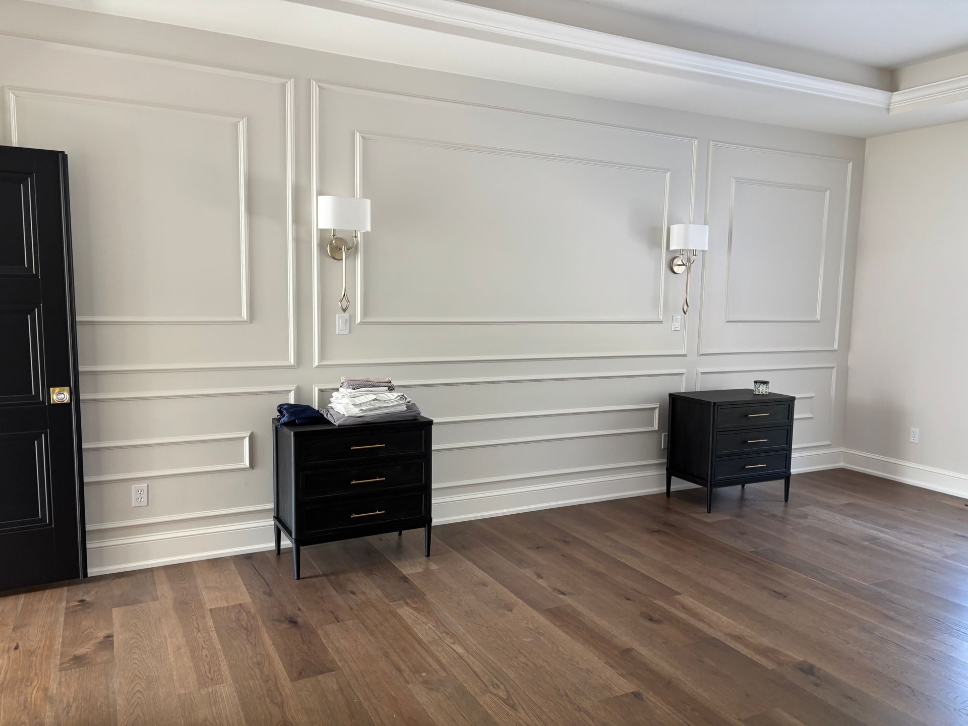 A bedroom with dark wooden floors and a wall with decorative white paneling, two black nightstands, and two wall sconces.