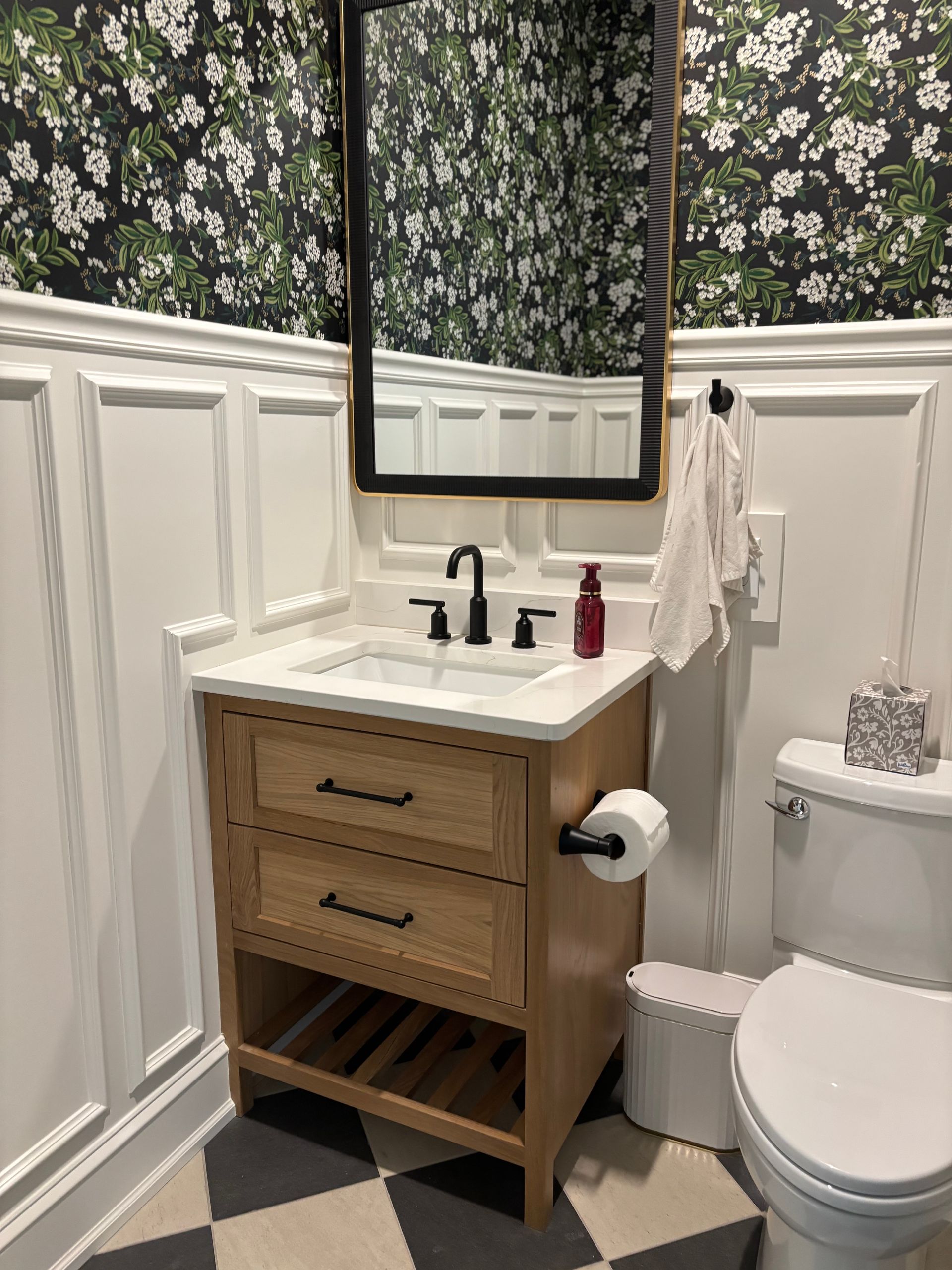 Bathroom with wooden vanity, toilet, floral wallpaper, and checkerboard floor.