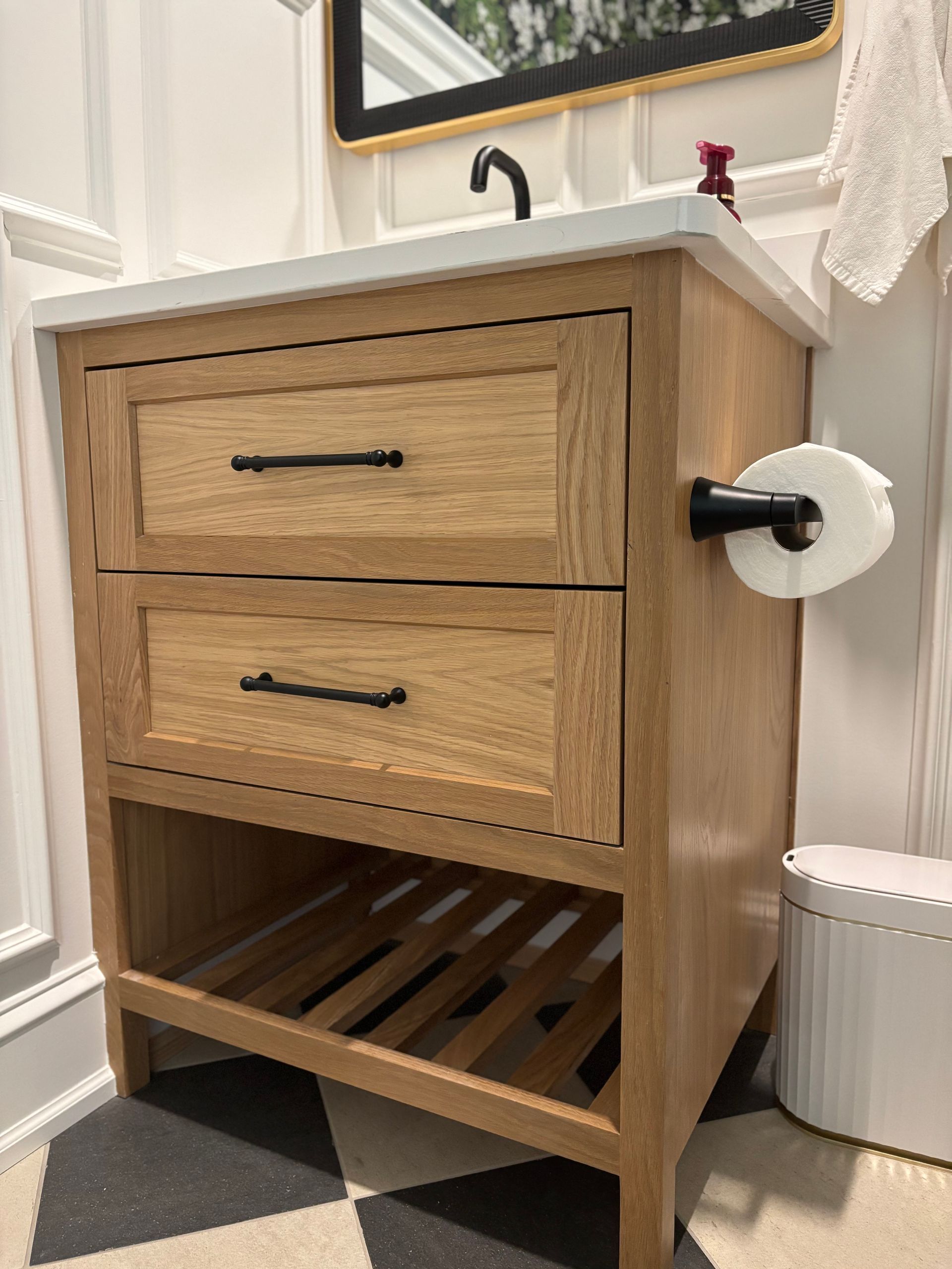 Wooden bathroom vanity with white countertop, two drawers, and open bottom shelf; black and white tiled floor.