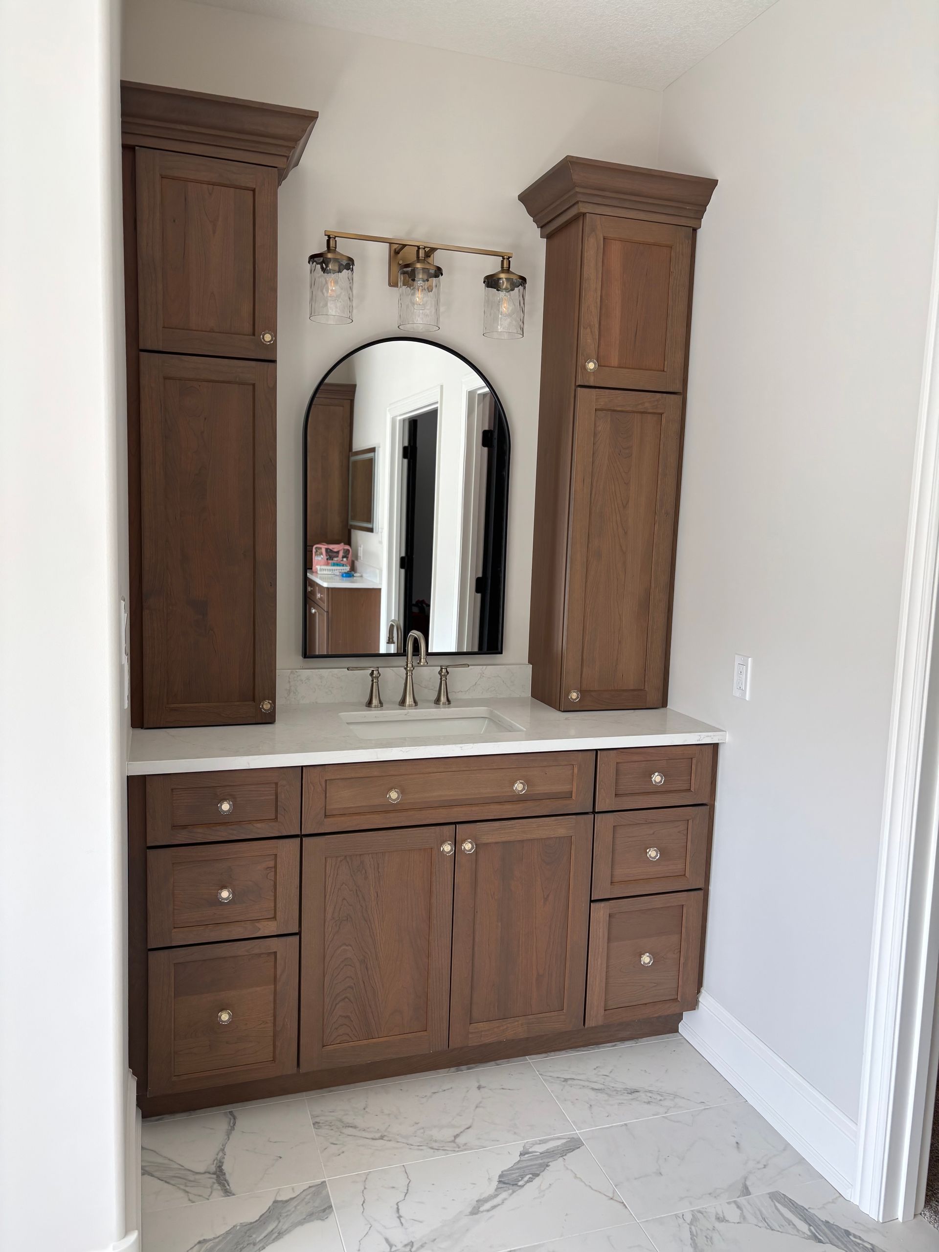 Bathroom vanity with brown cabinetry, marble countertop, and arched mirror.
