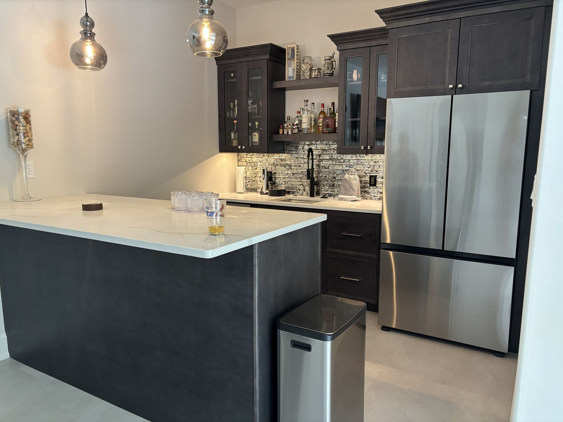 A modern home bar with dark cabinets, a stainless steel refrigerator, and a white countertop.