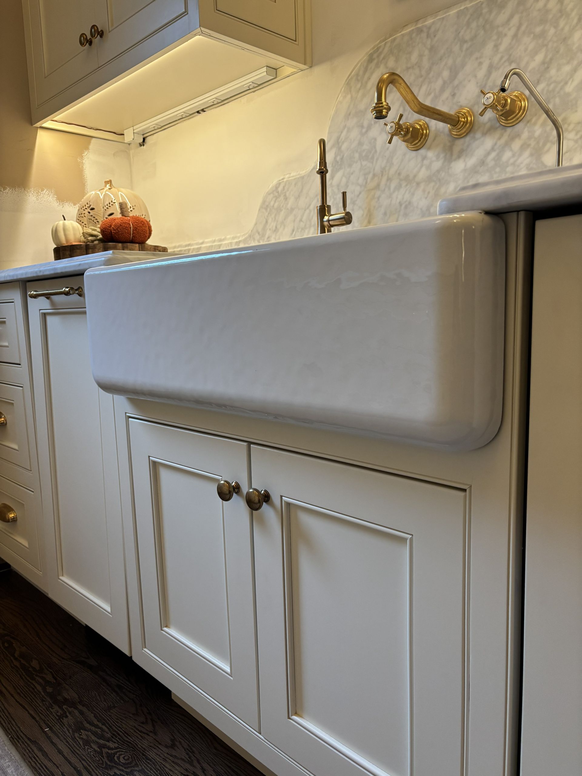 White farmhouse sink in a kitchen with brass fixtures and light-colored cabinets.